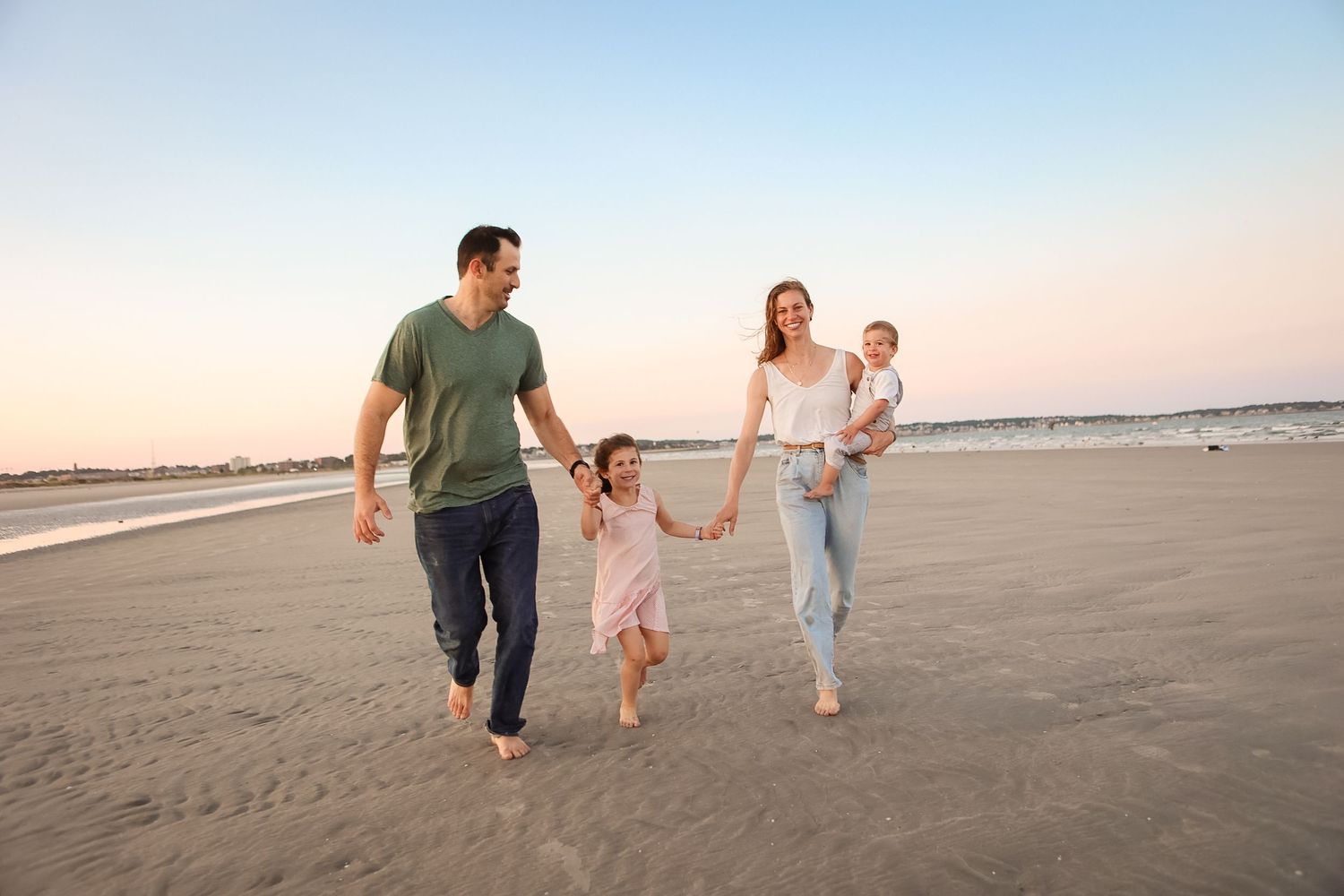 Happy family of four walking barefoot on sandy beach at sunset, holding hands.