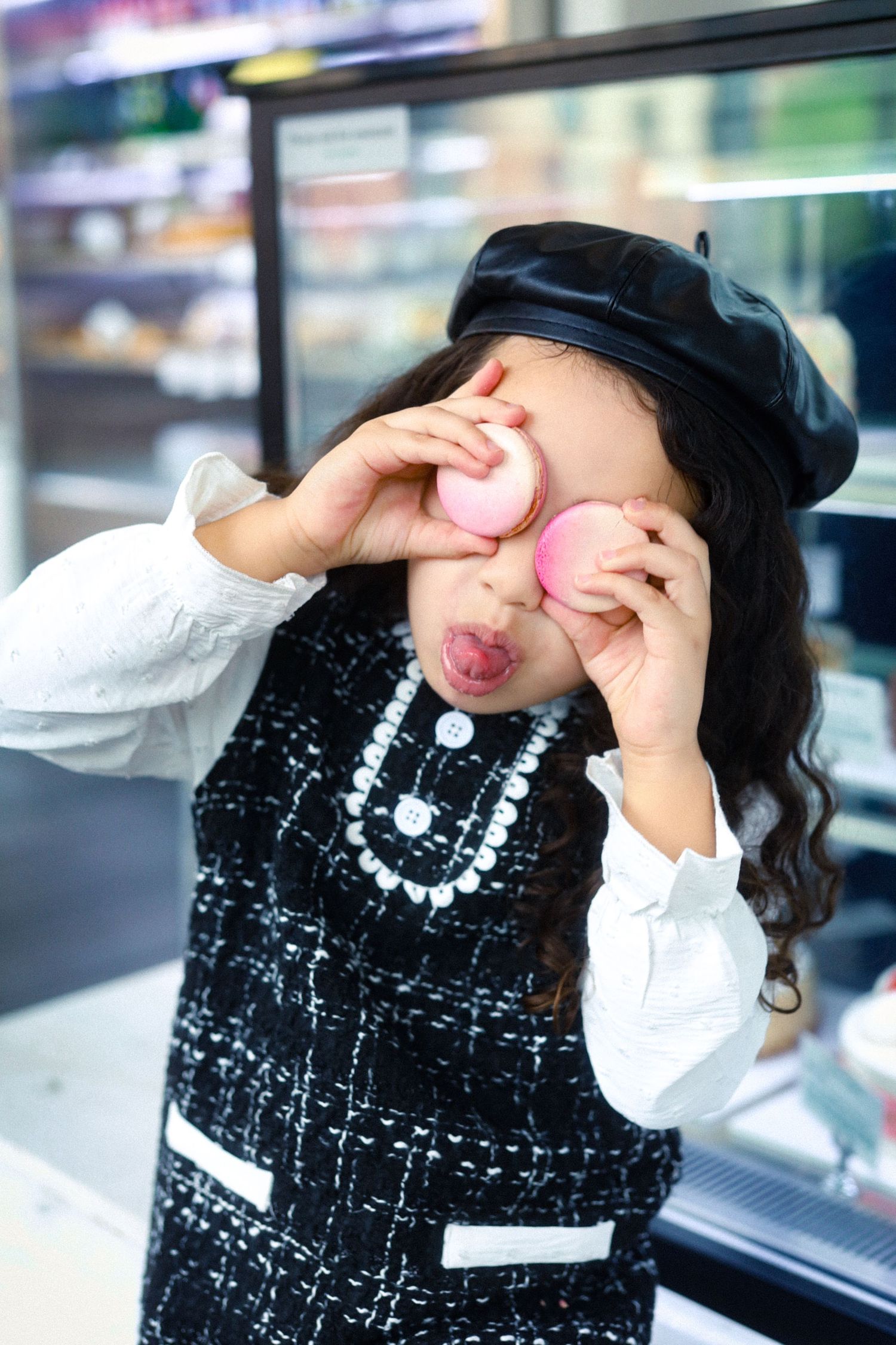 Playful young girl in black beret and tweed outfit holding macarons over her eyes.