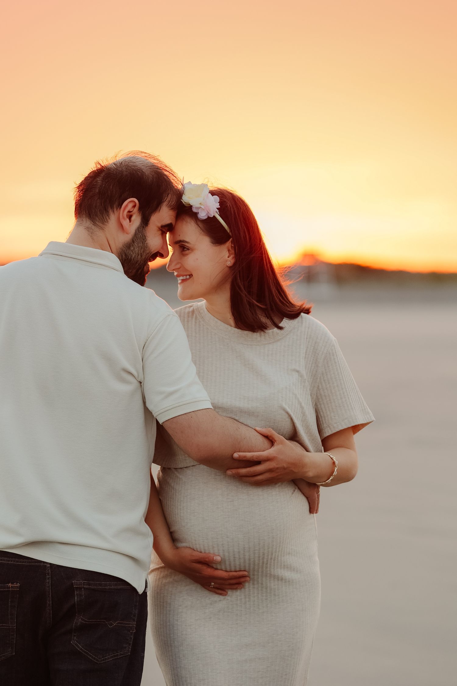 Couple sharing an intimate moment at sunset, pregnant woman in white dress with flower crown holding her baby bump.