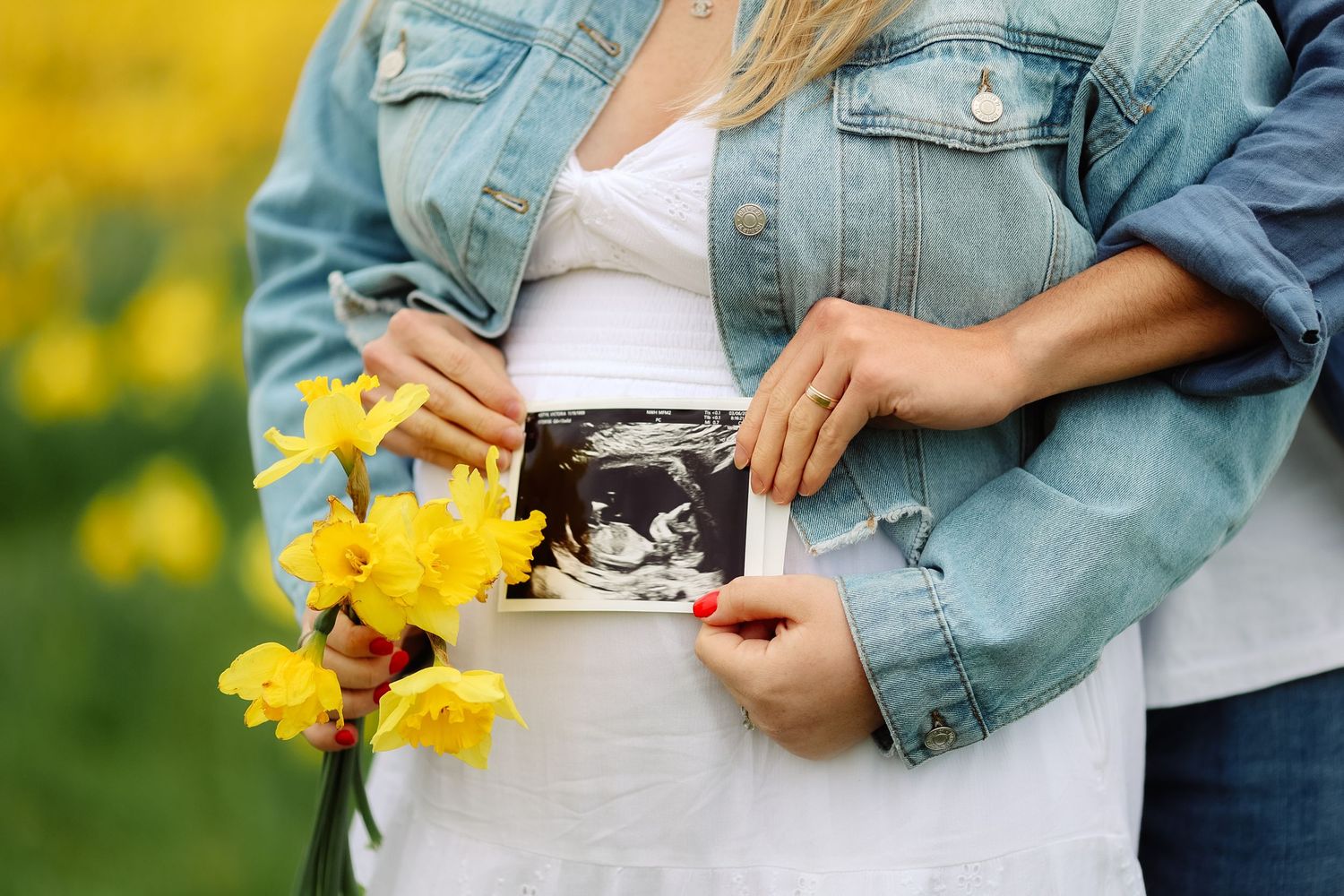 Couple holding ultrasound scan and yellow daffodils, announcing pregnancy in a blooming flower field.