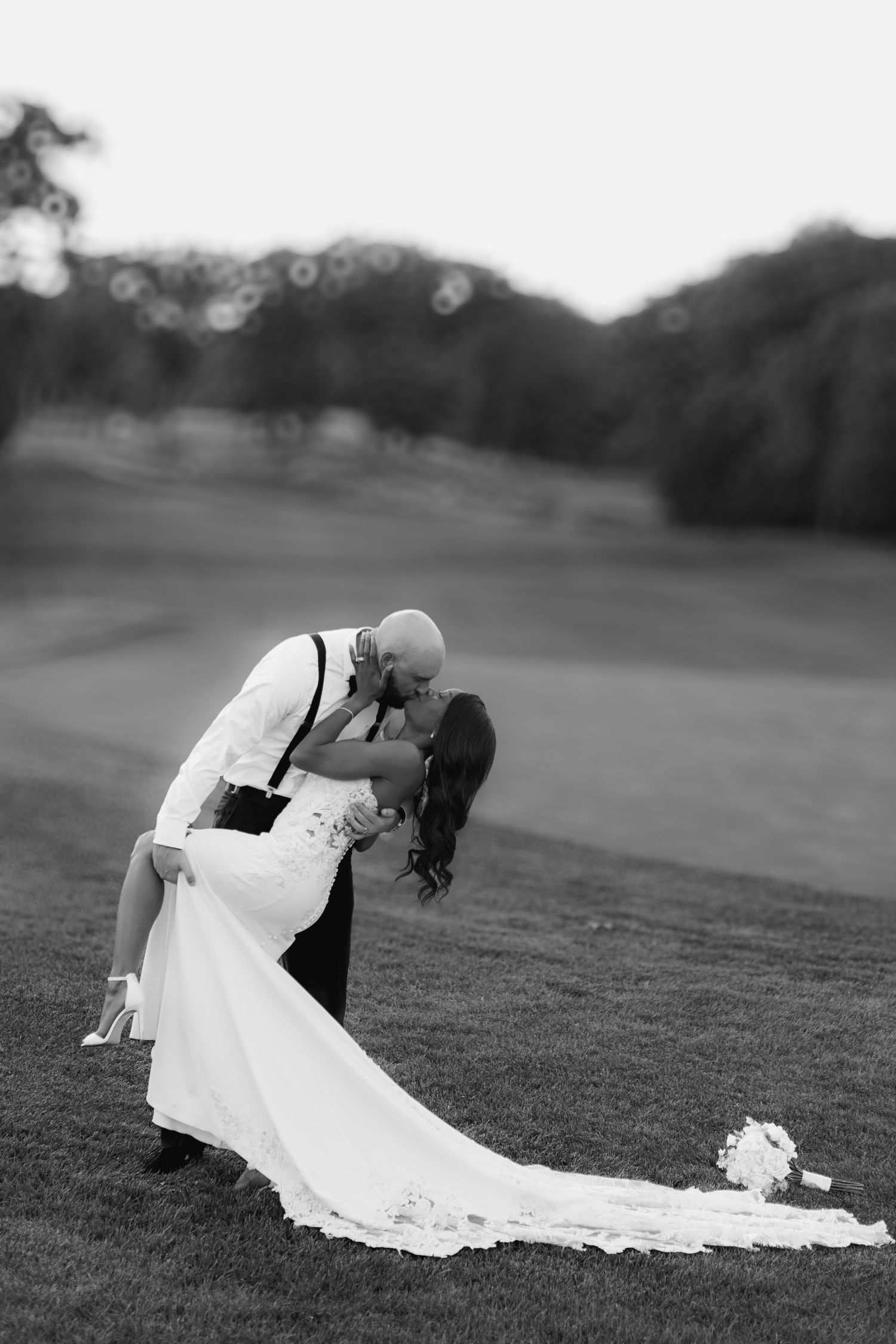 Black and white photo of groom dipping bride for a kiss on a golf course, her bouquet resting on the grass nearby.
