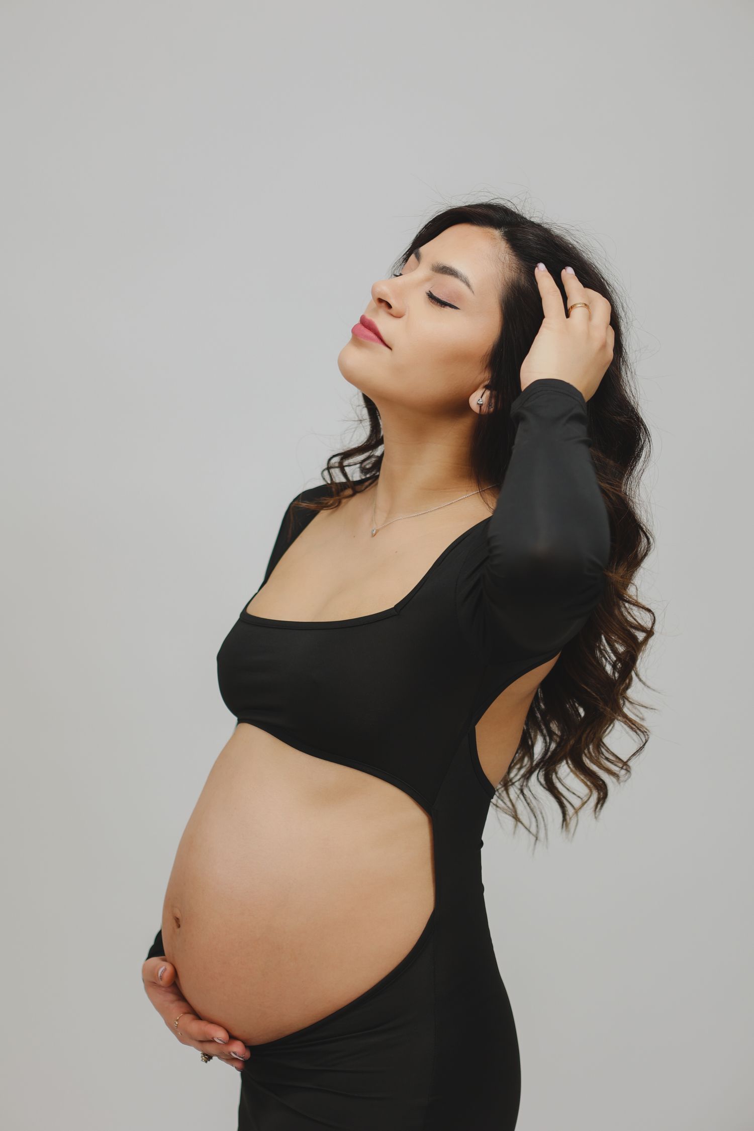 Elegant pregnant woman in black cutout dress cradling baby bump against light gray background.