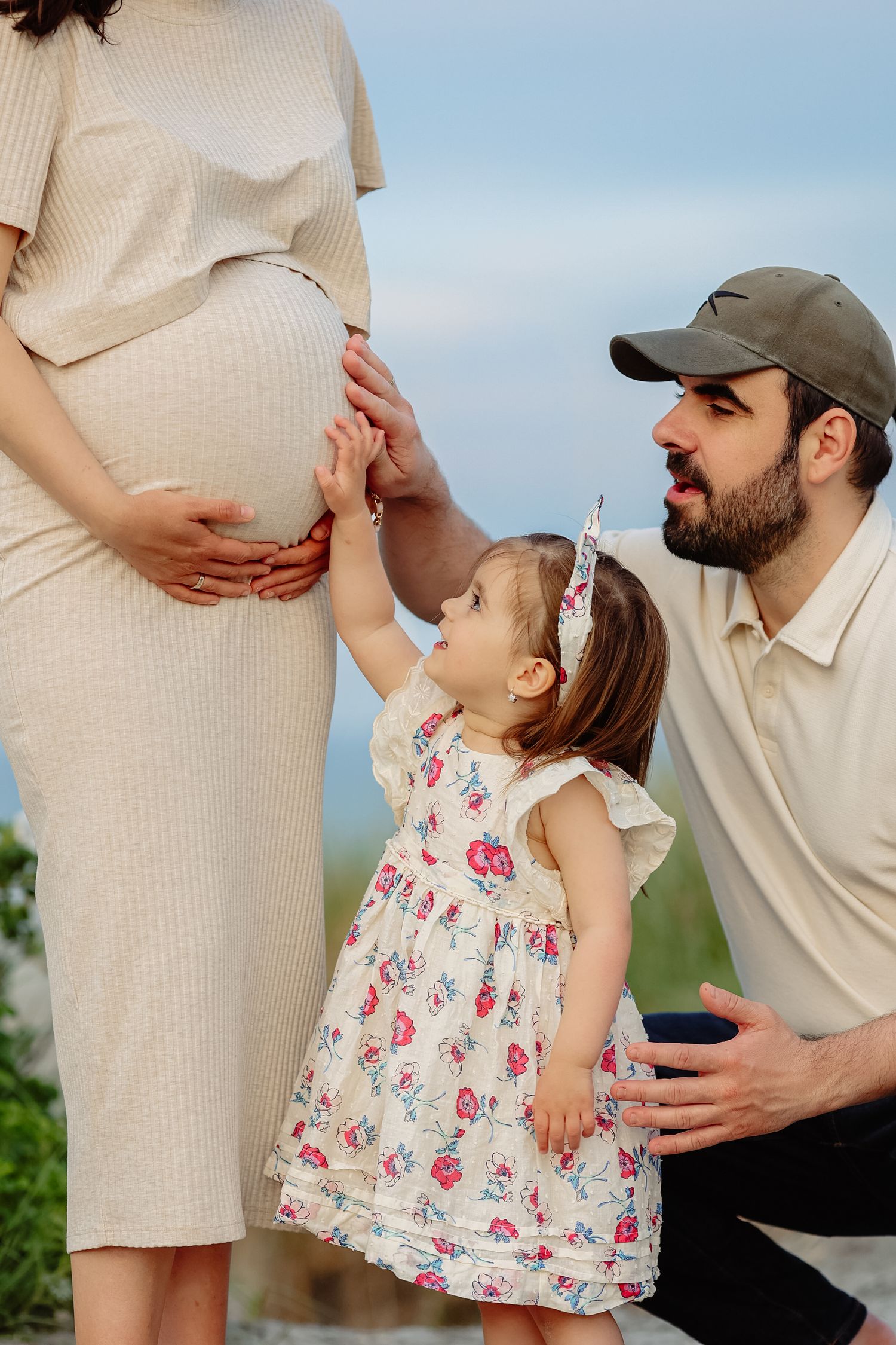 Bearded father kneels beside toddler daughter as she touches pregnant mothers baby bump outdoors.