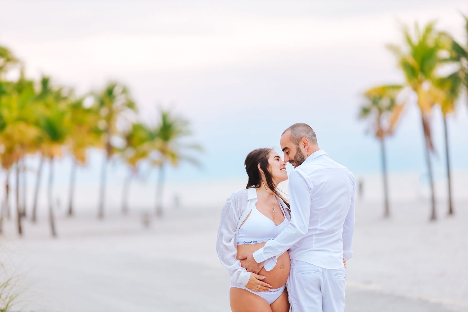 Romantic pregnant couple in white outfits embracing on tropical beach with palm trees at sunset.