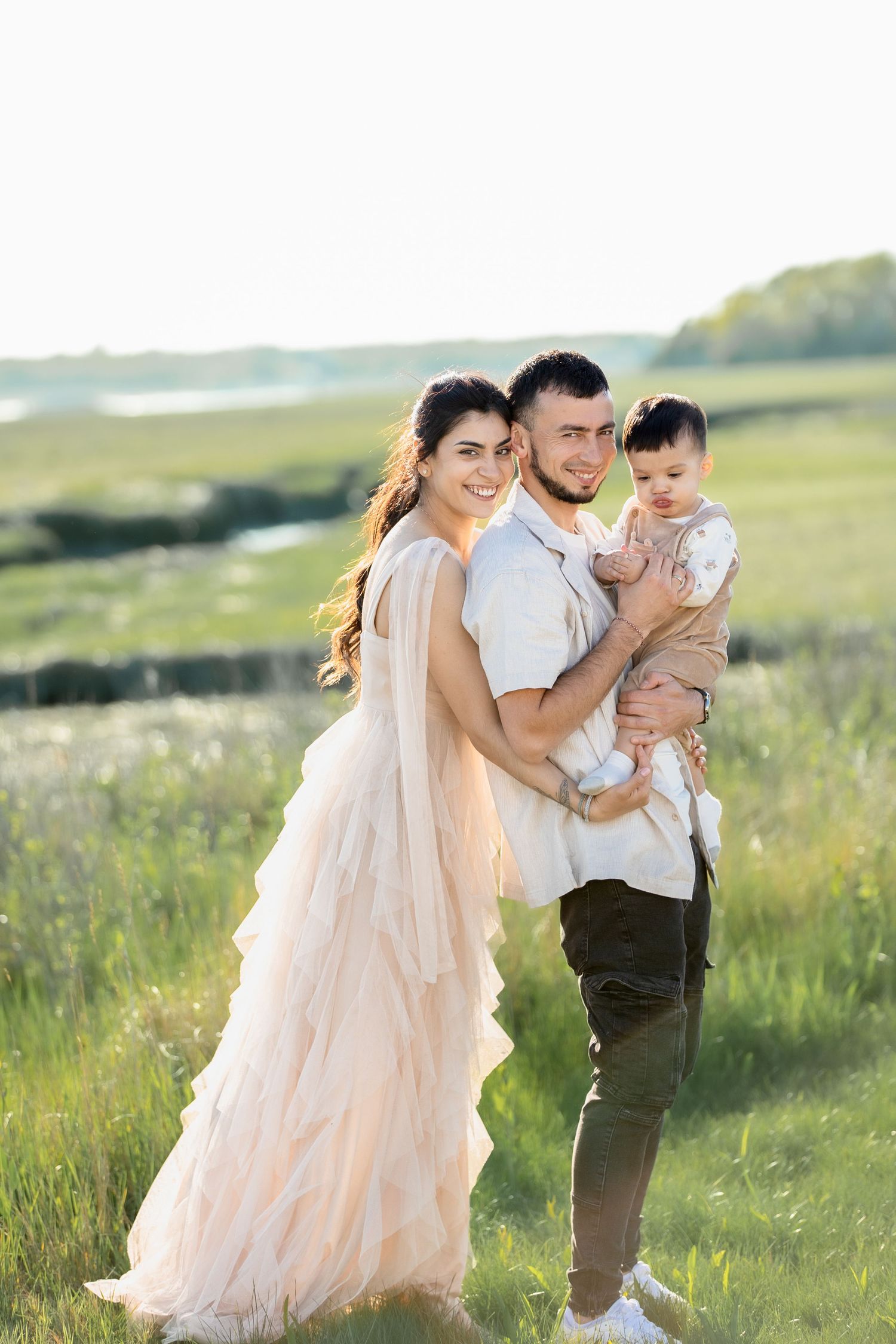Happy family with baby posing in sunlit green meadow together.