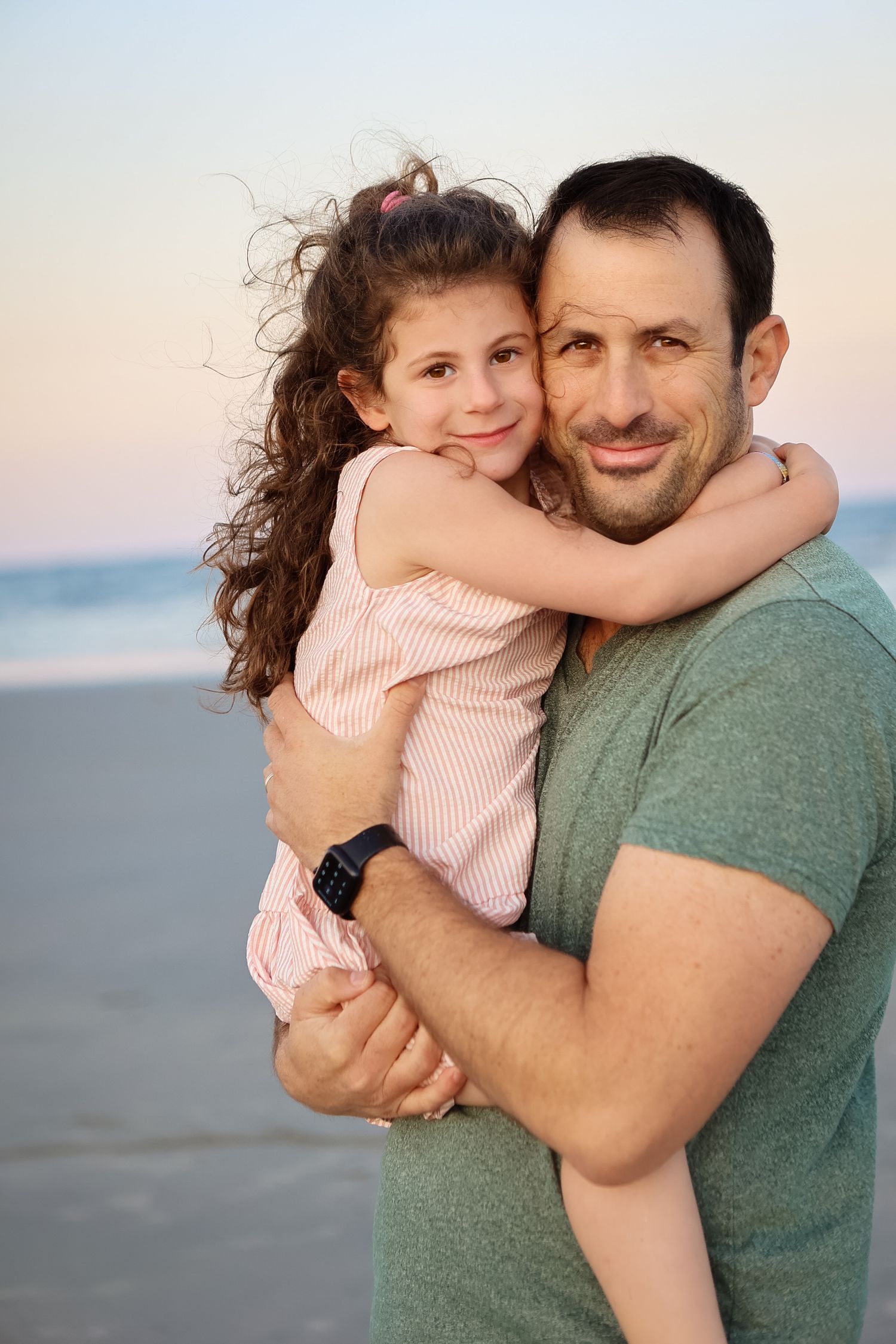 Loving father holding young curly-haired daughter in pink dress on beach at dusk.