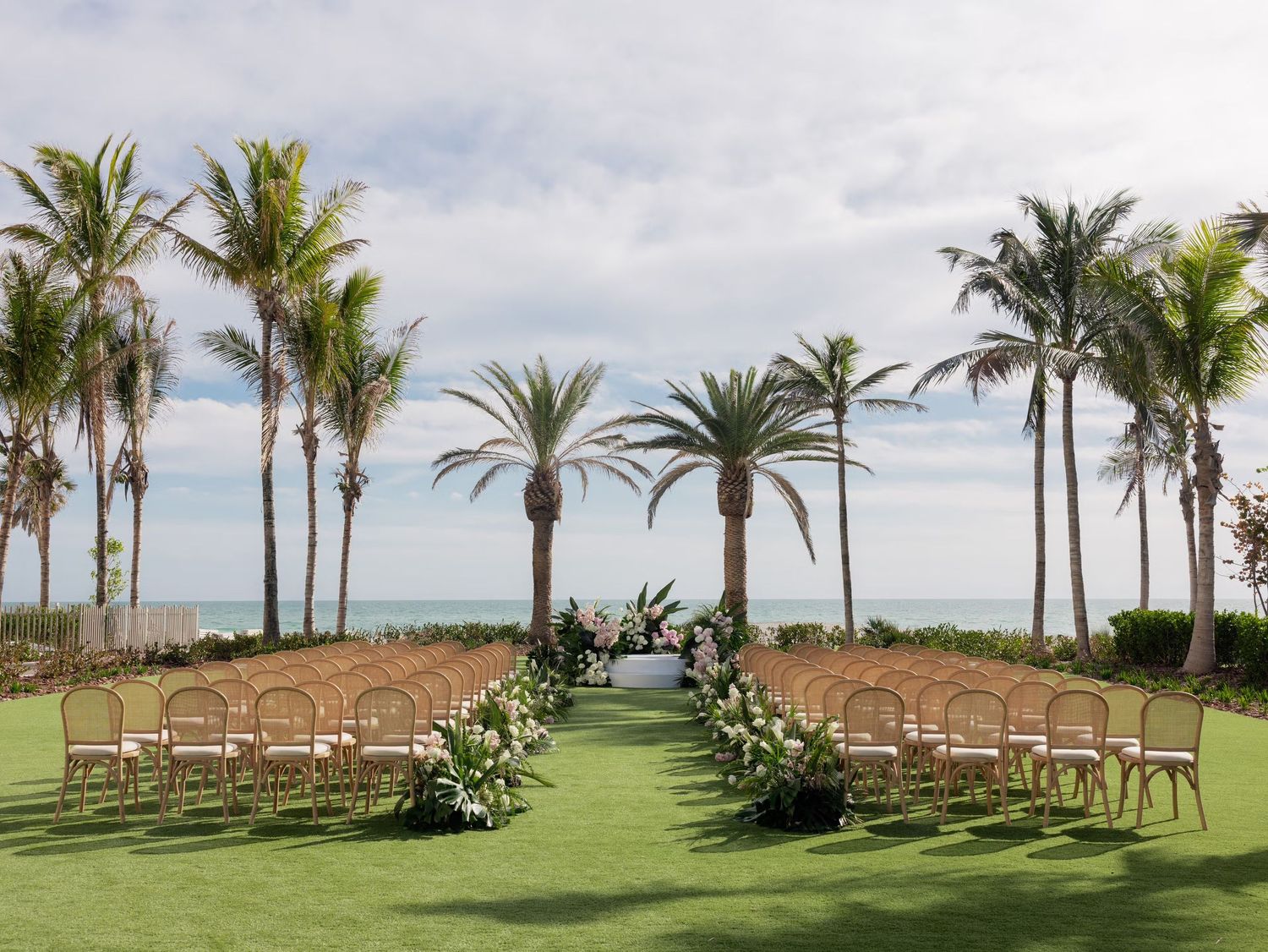 Outdoor beach wedding ceremony setup with rattan chairs, floral arrangements, and tall palm trees overlooking the ocean.