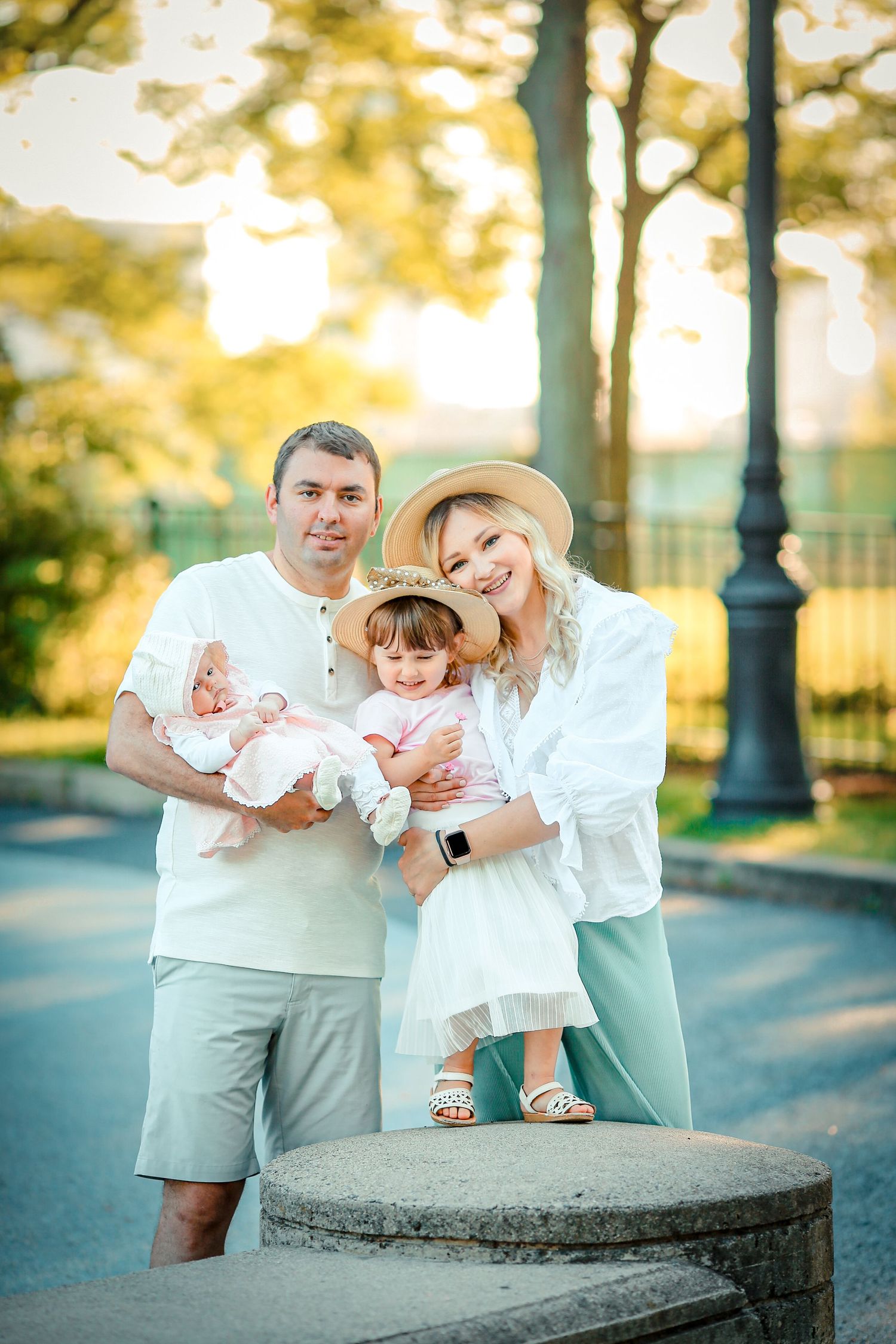 Happy family of four posing outdoors at sunset, parents holding two young children in white outfits.