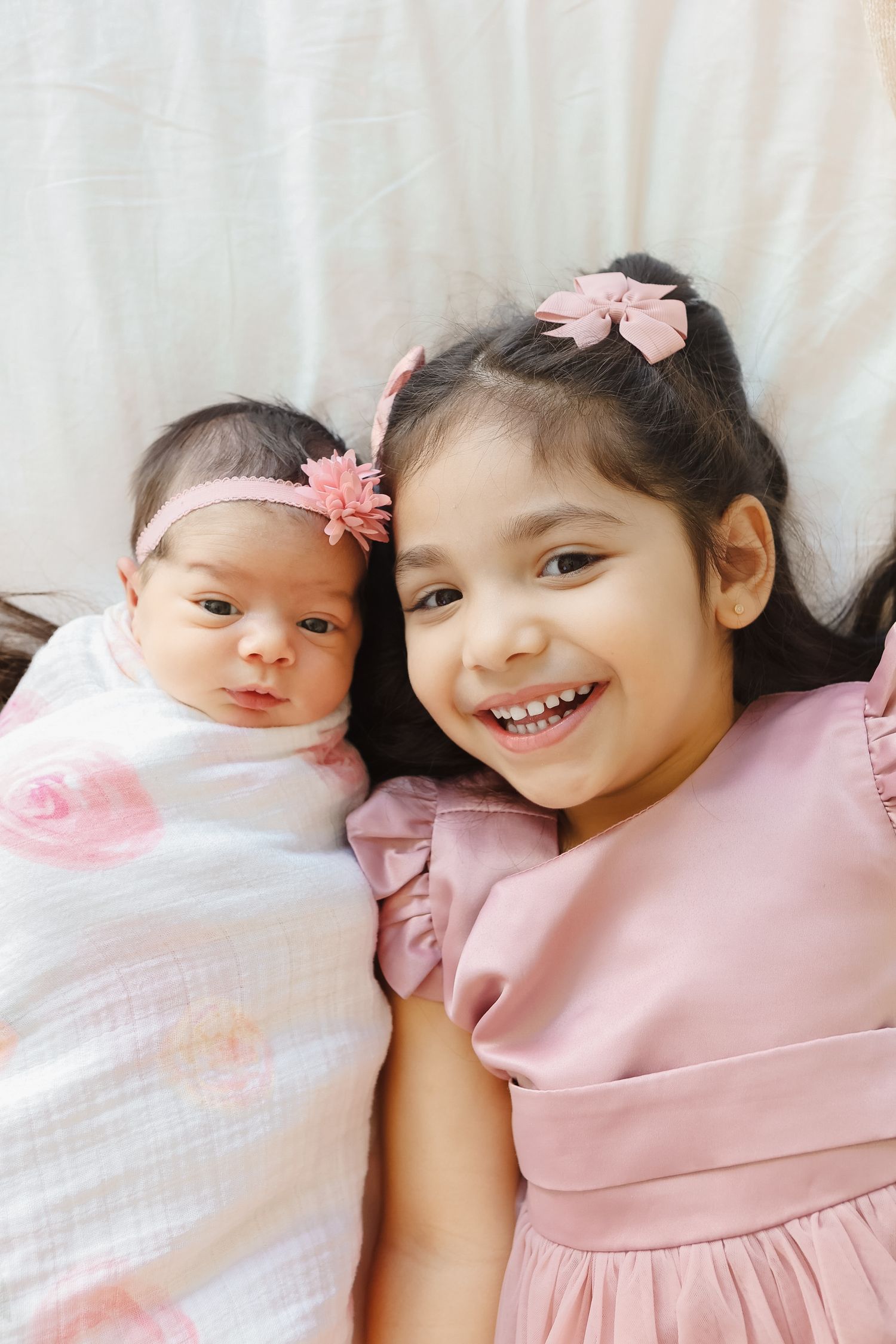 Smiling big sister in pink dress lying next to newborn in floral swaddle.