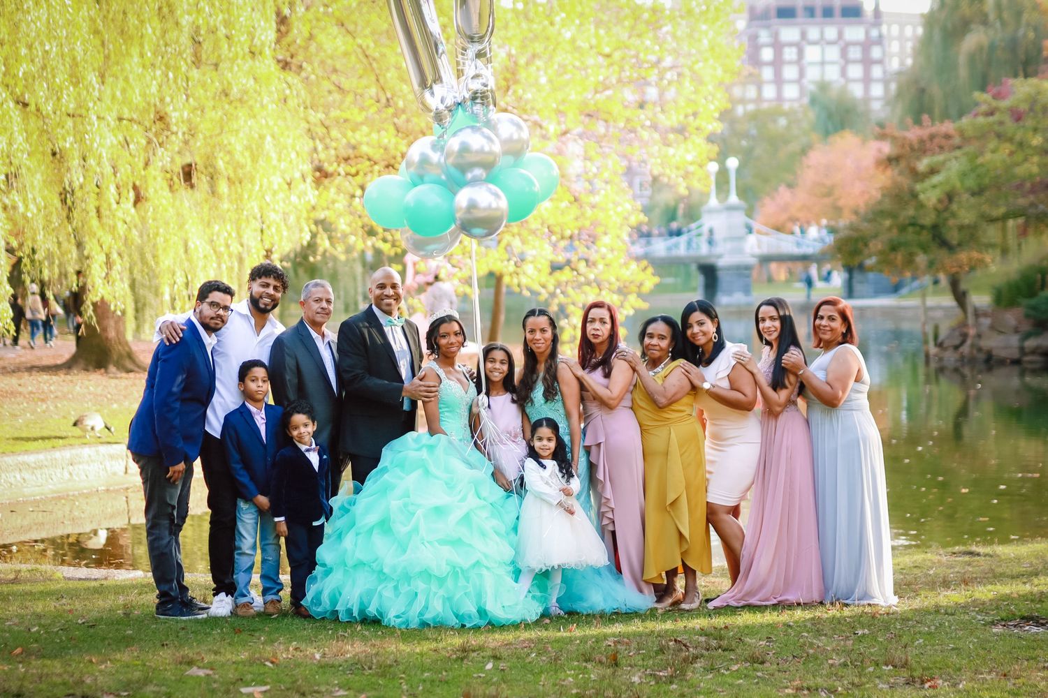 Large extended family group celebrating a quinceañera outdoors near a pond with teal balloons.