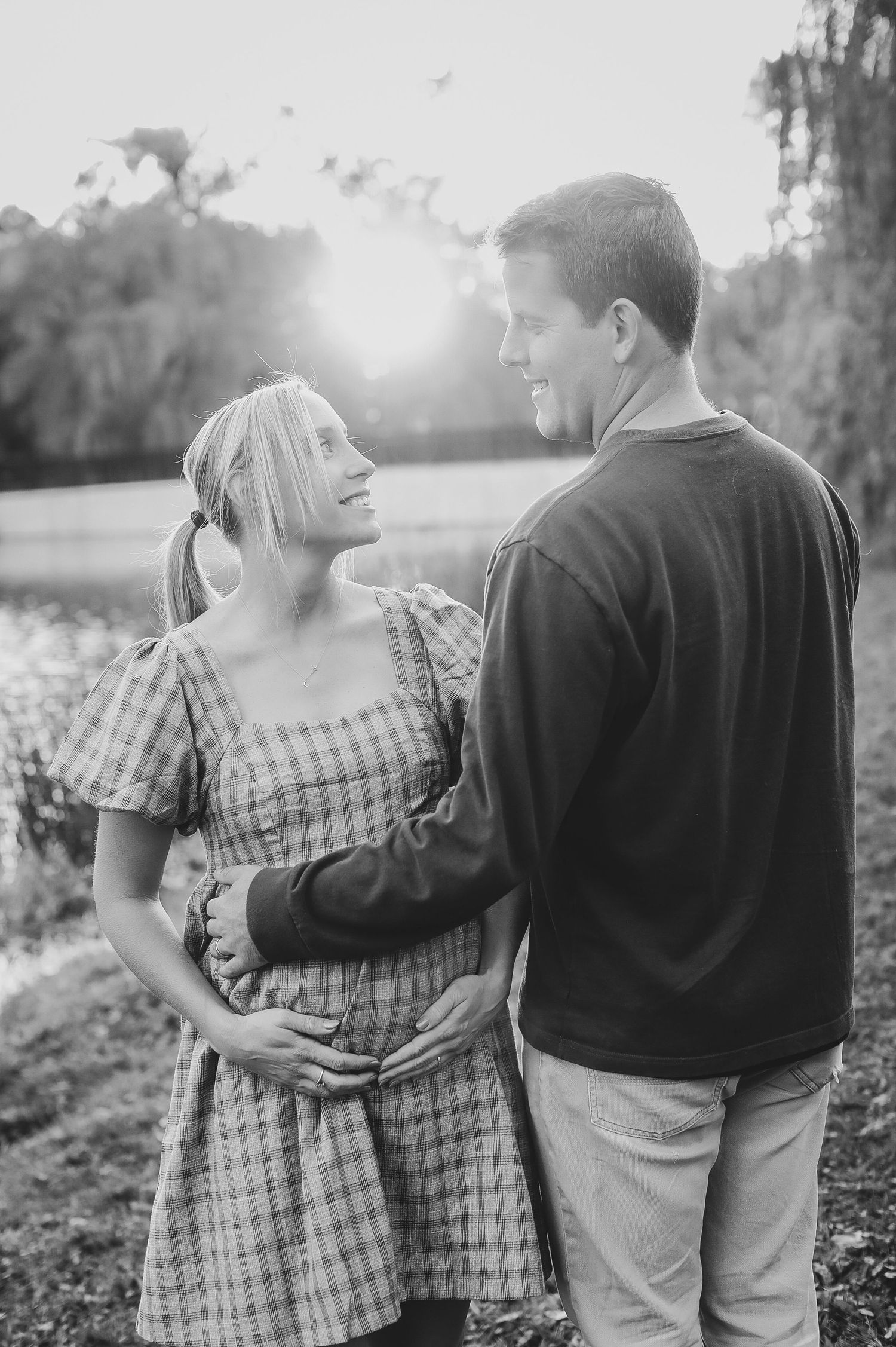 Black and white maternity photo of smiling couple embracing at golden hour near a sunlit lake.
