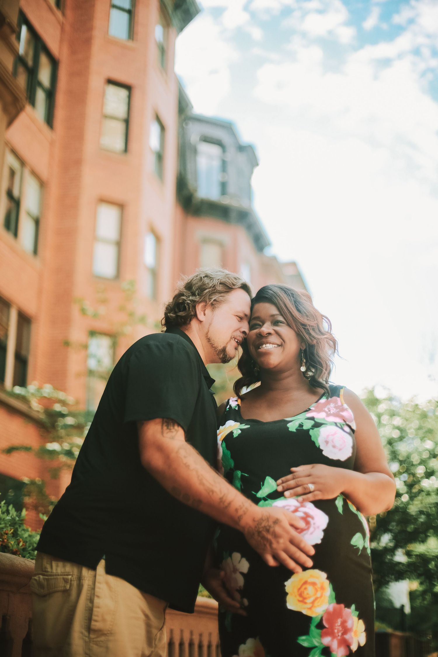 Happy interracial couple embracing outdoors, pregnant woman in floral dress smiling near brick buildings.