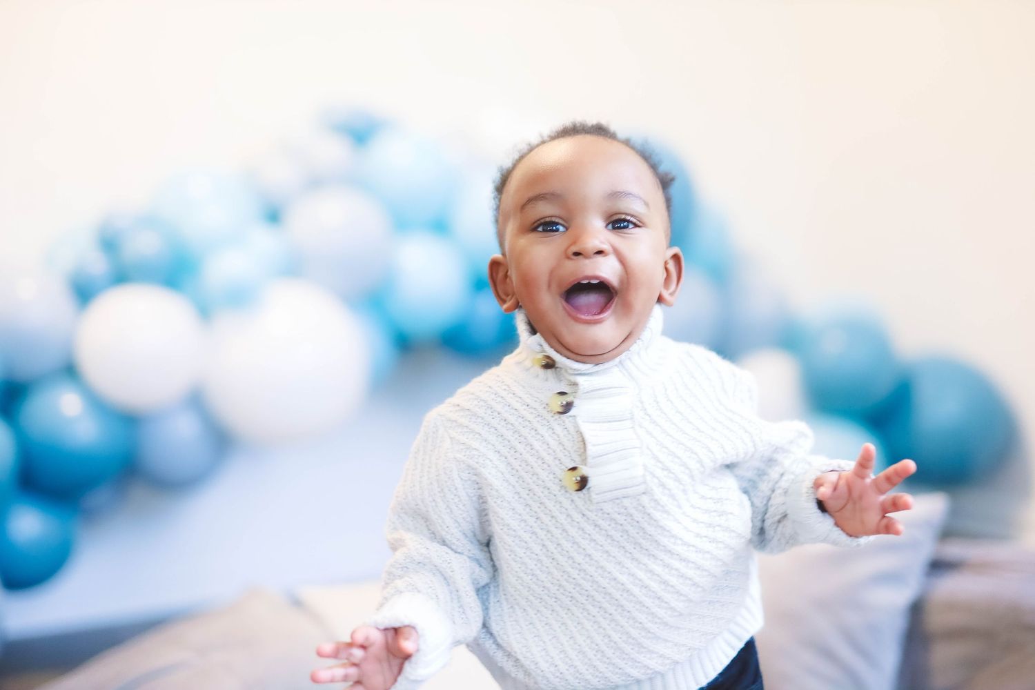 Joyful baby boy in white sweater laughing with blue and white balloon garland behind him.