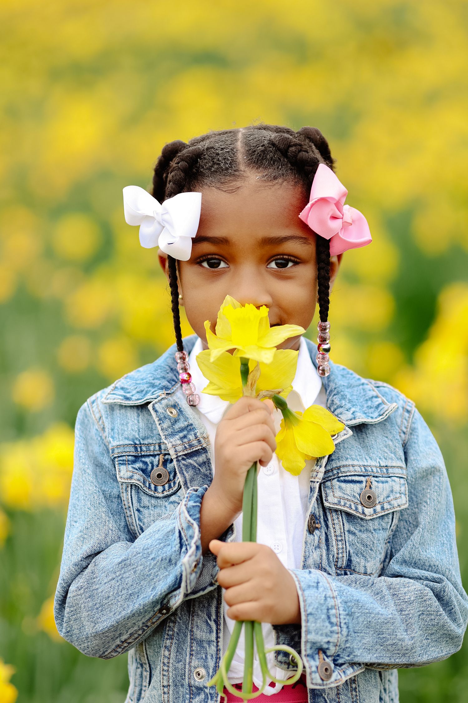 Young Black girl smelling yellow daffodils in blooming flower field, wearing denim jacket and hair bows.