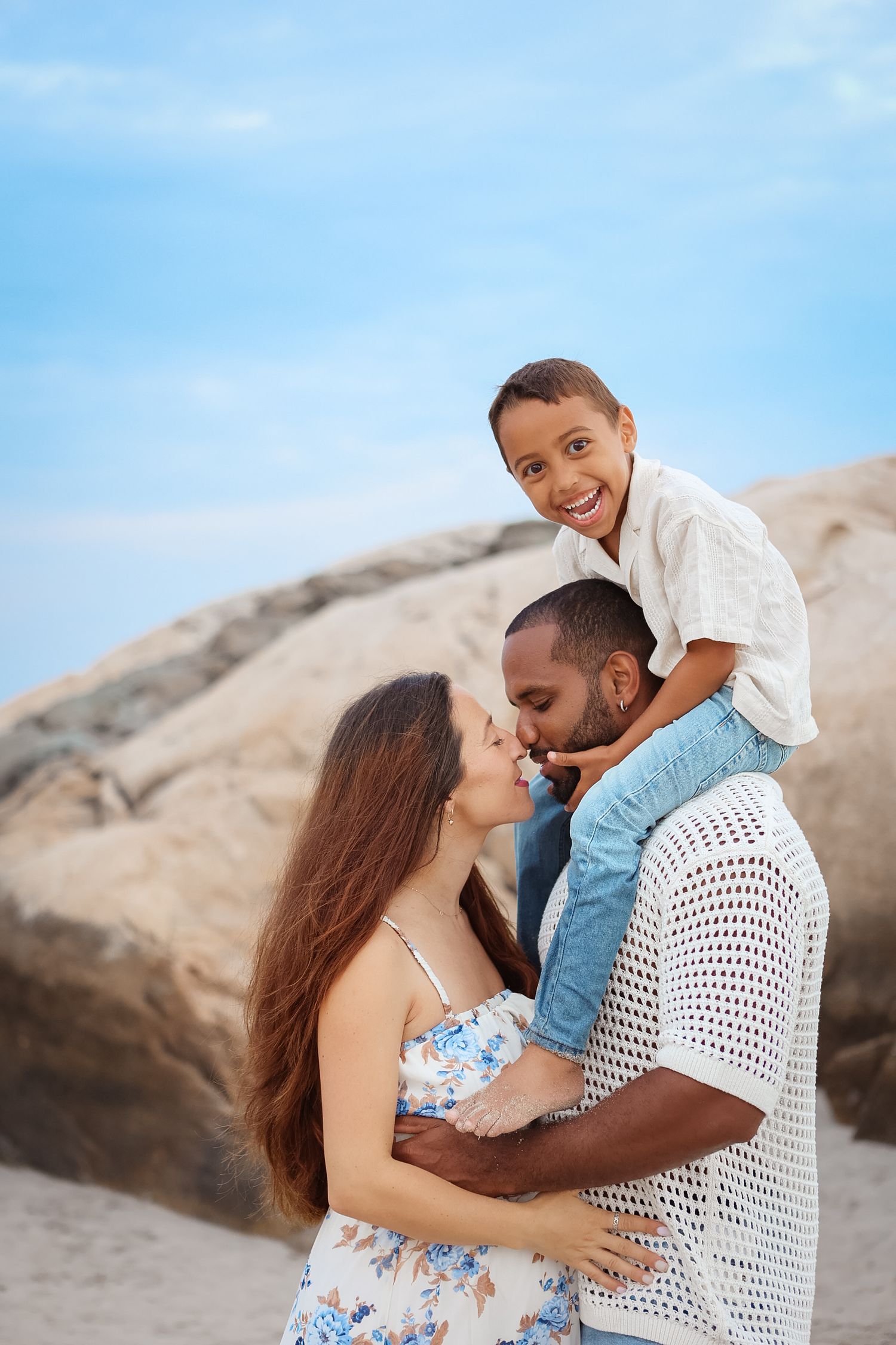 Happy interracial family at beach with son sitting on fathers shoulders as parents kiss.