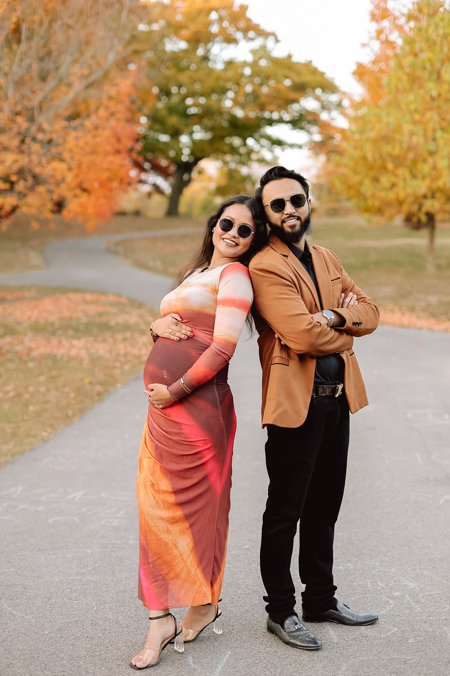 Stylish pregnant couple posing back-to-back in colorful fall park during maternity photoshoot.