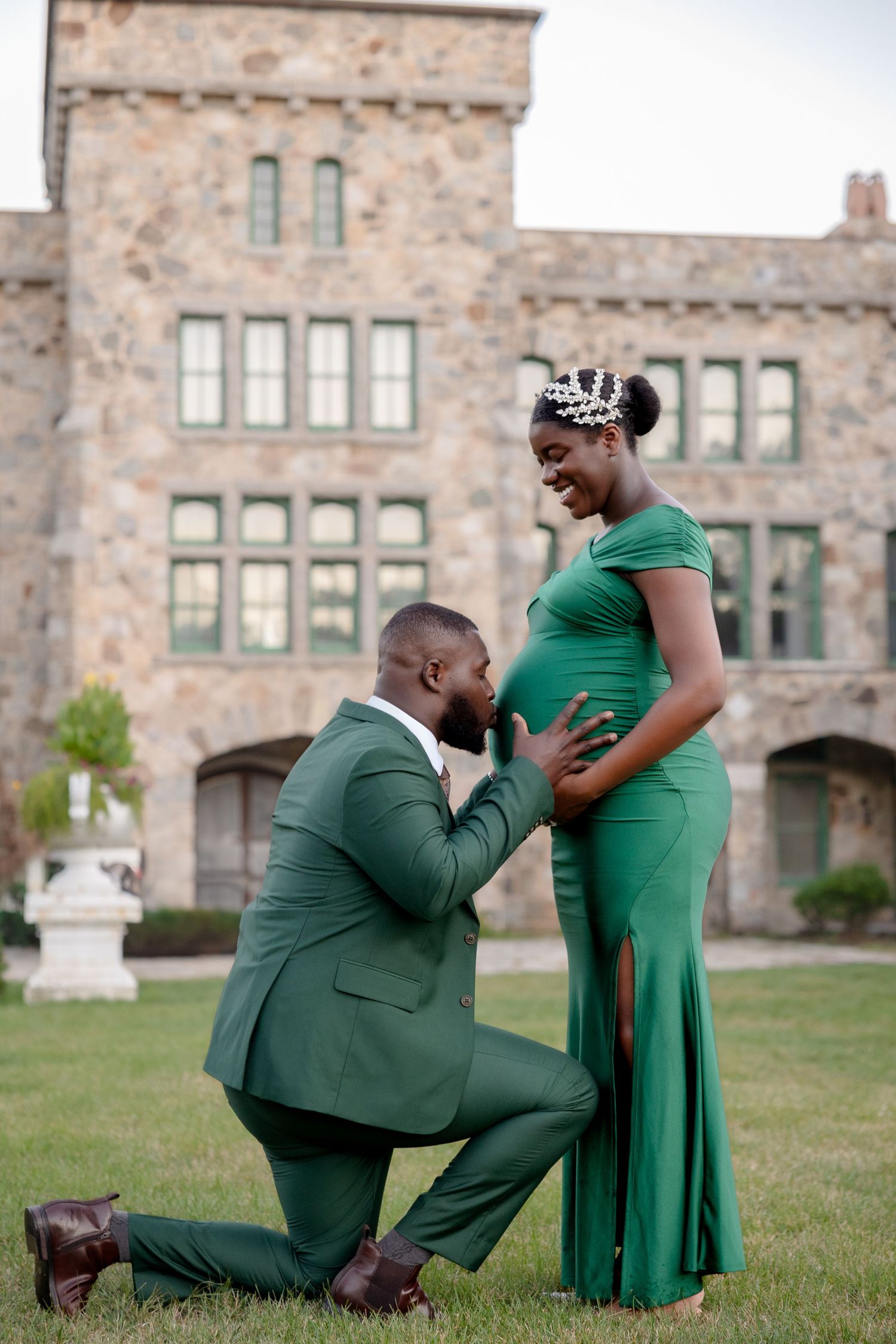Man in green suit kneeling to kiss pregnant womans belly during outdoor maternity photoshoot near stone building.