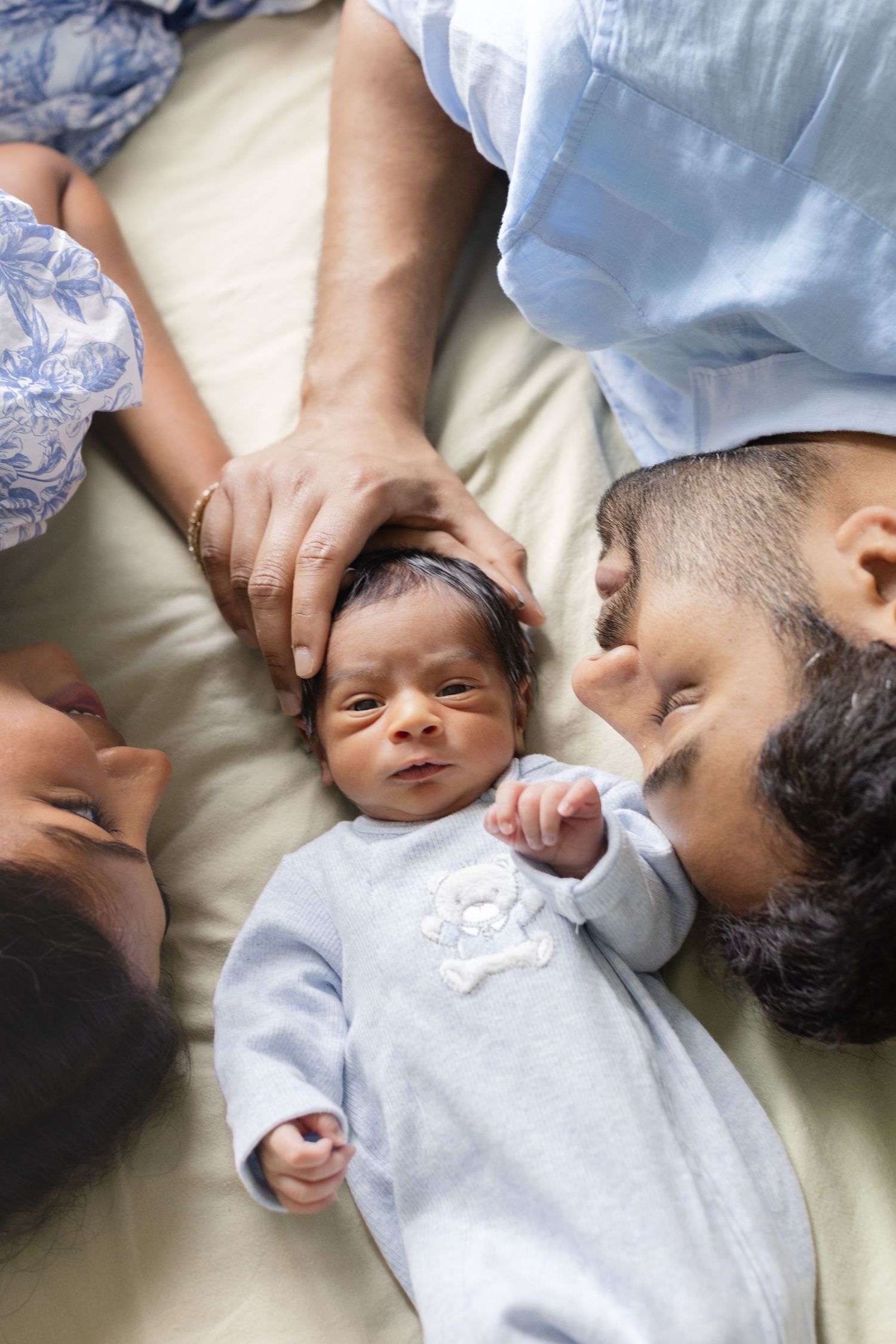 Top view of parents lying with newborn baby in blue onesie on bed.