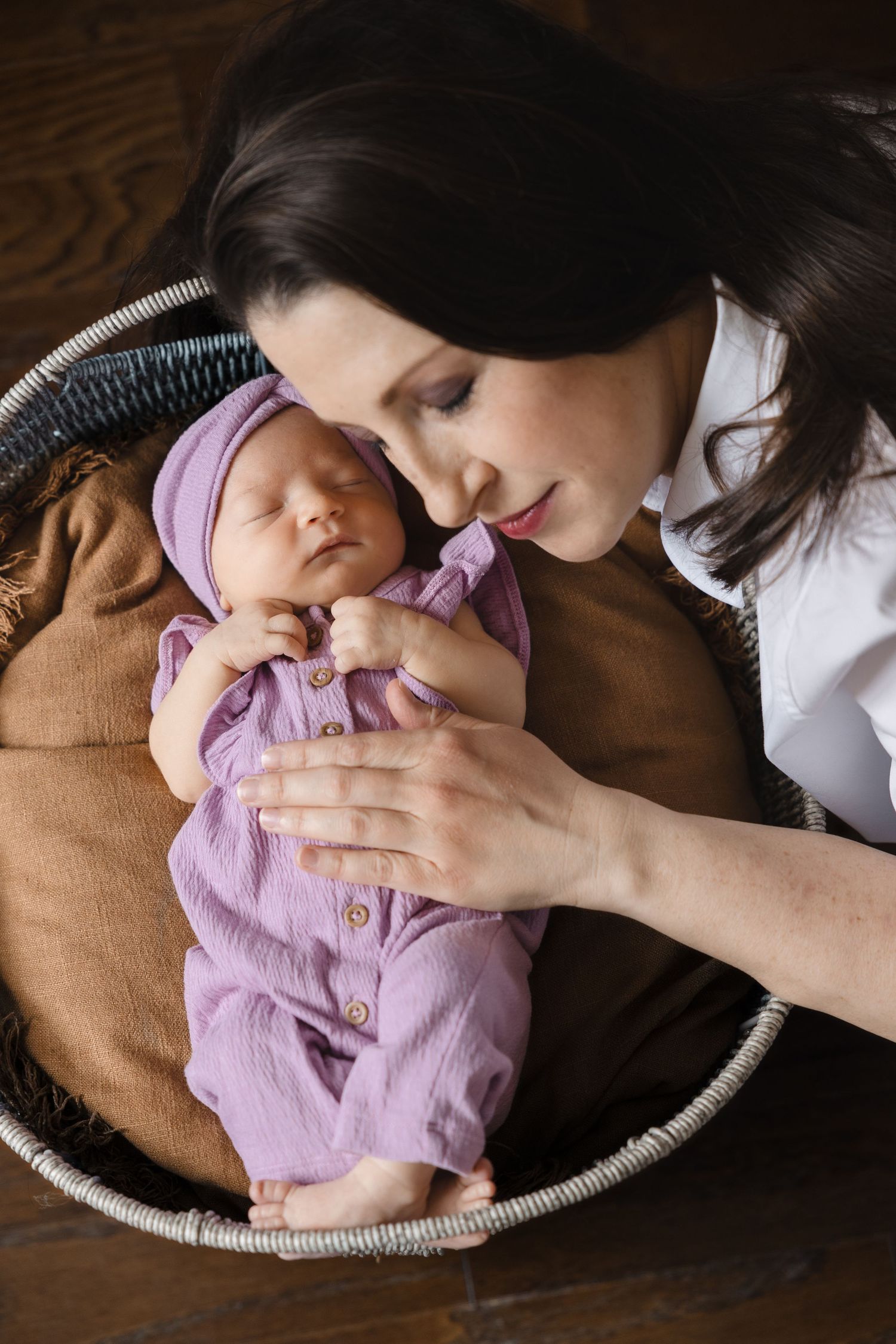 Loving mother leans over sleeping newborn baby wrapped in purple swaddle inside wicker basket.