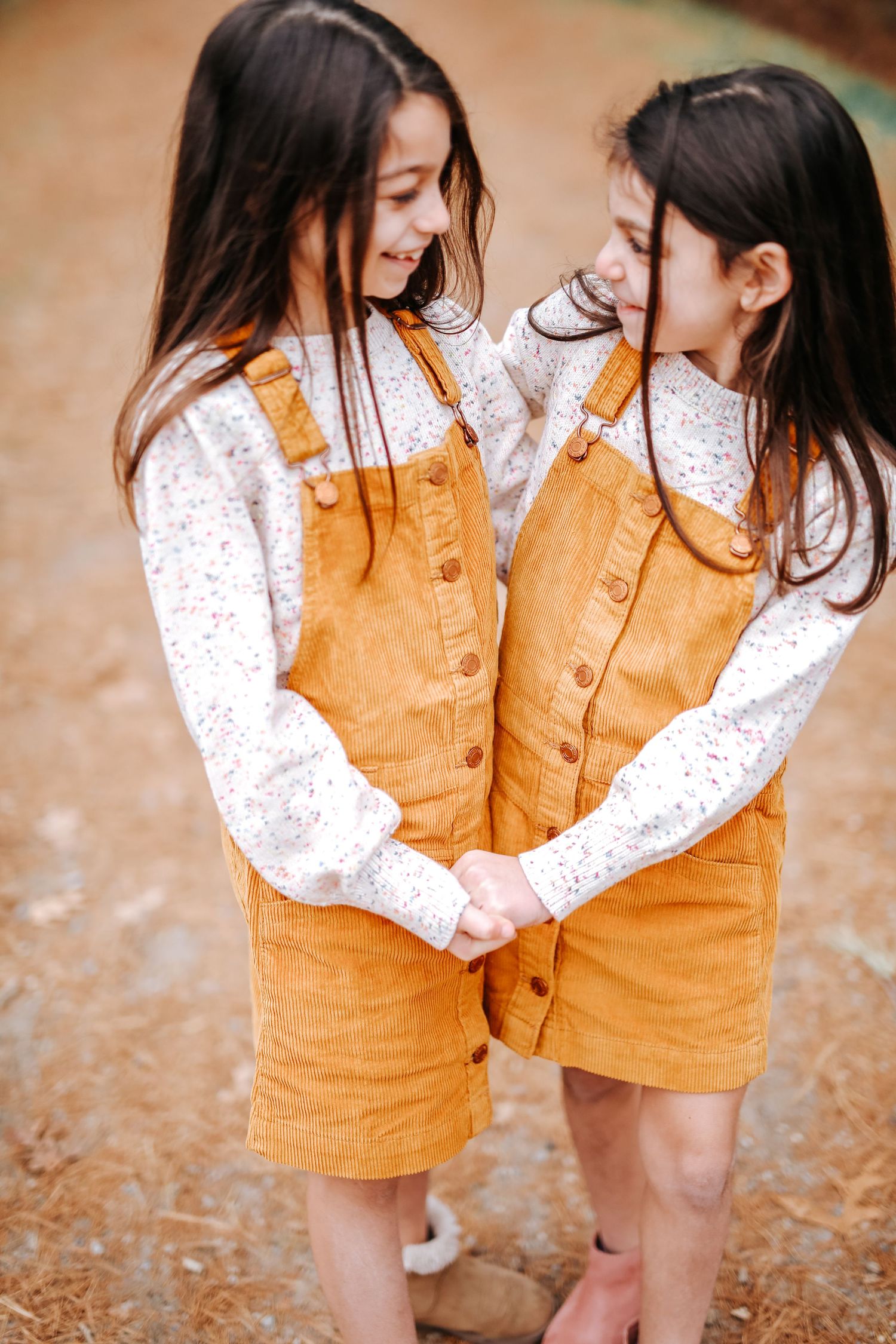 Twin girls in matching mustard corduroy overalls holding hands and smiling at each other outdoors.