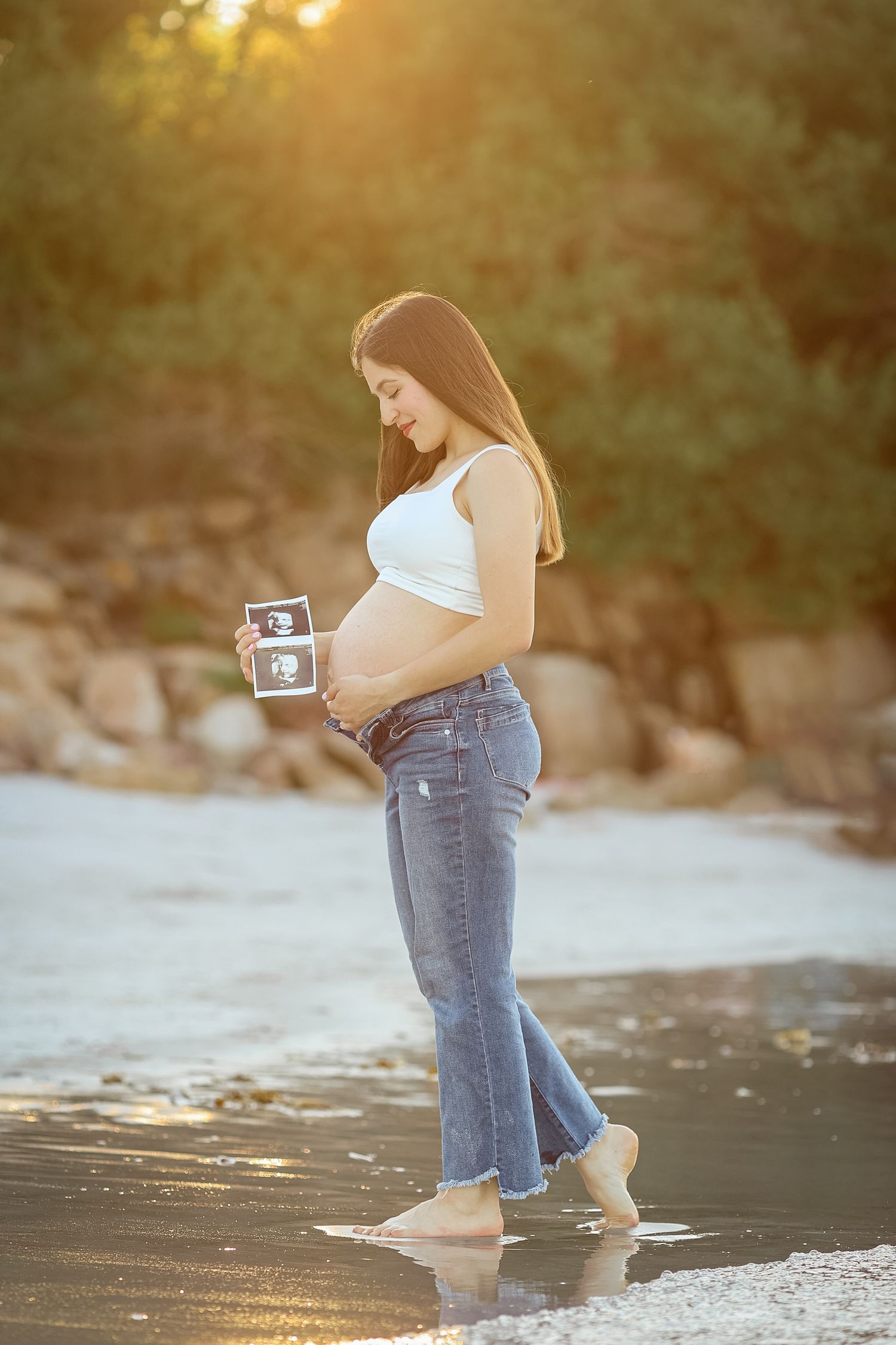 Pregnant woman in white crop top and jeans holds ultrasound photos while standing on a sunlit beach.