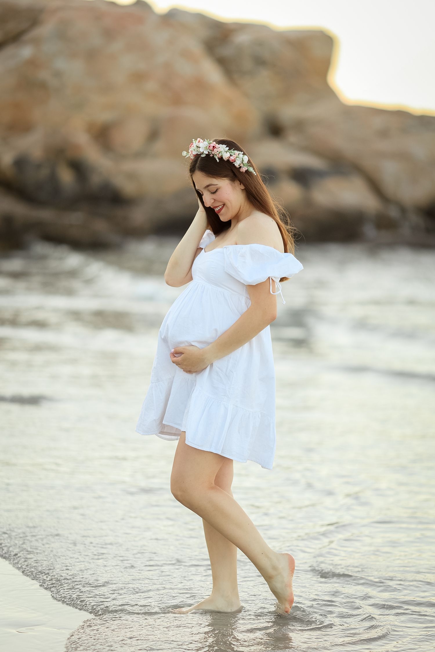 Pregnant woman in white off-shoulder dress and floral crown wades barefoot in ocean waves at sunset.