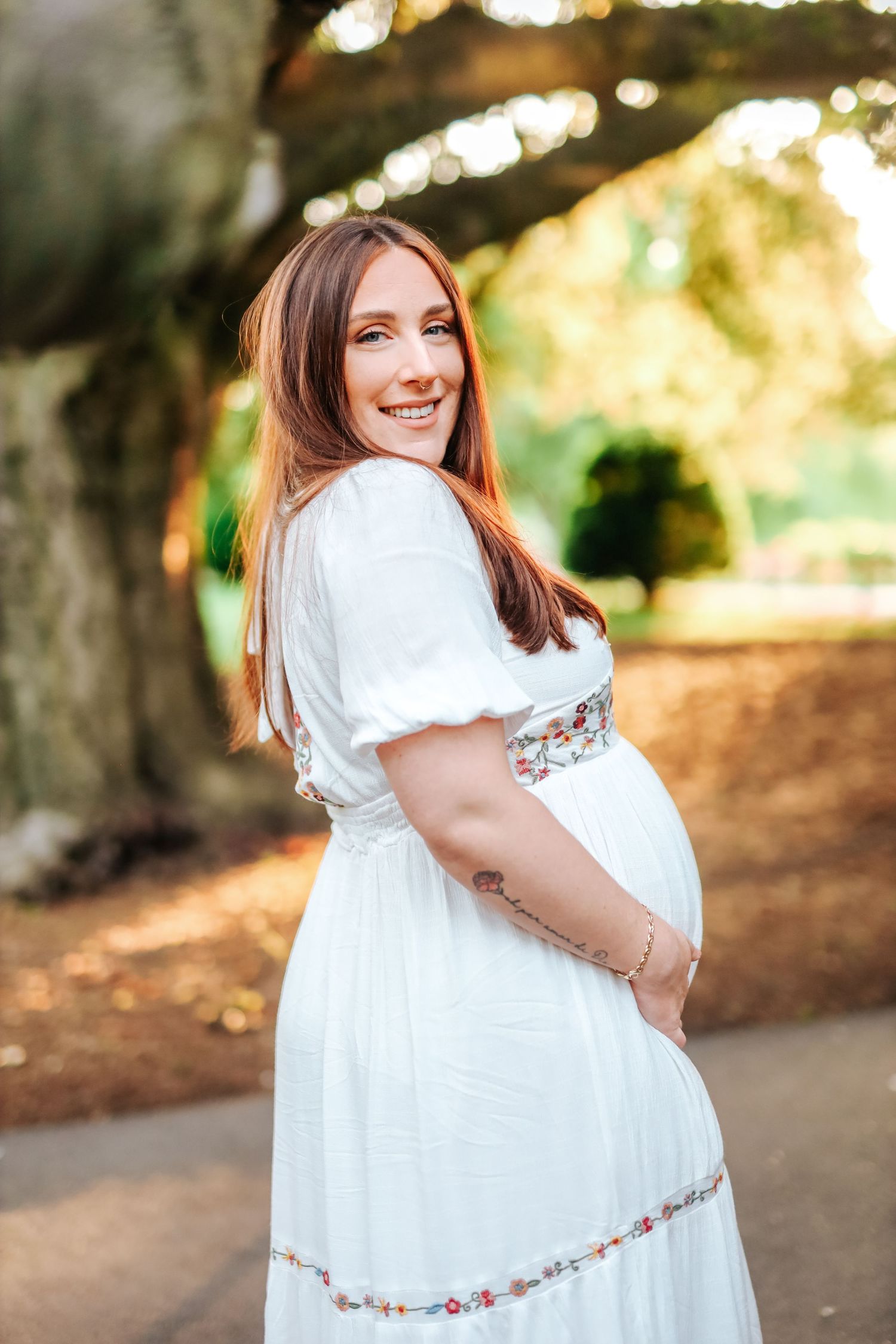 Smiling pregnant woman in white embroidered dress posing in sunlit park during maternity shoot.
