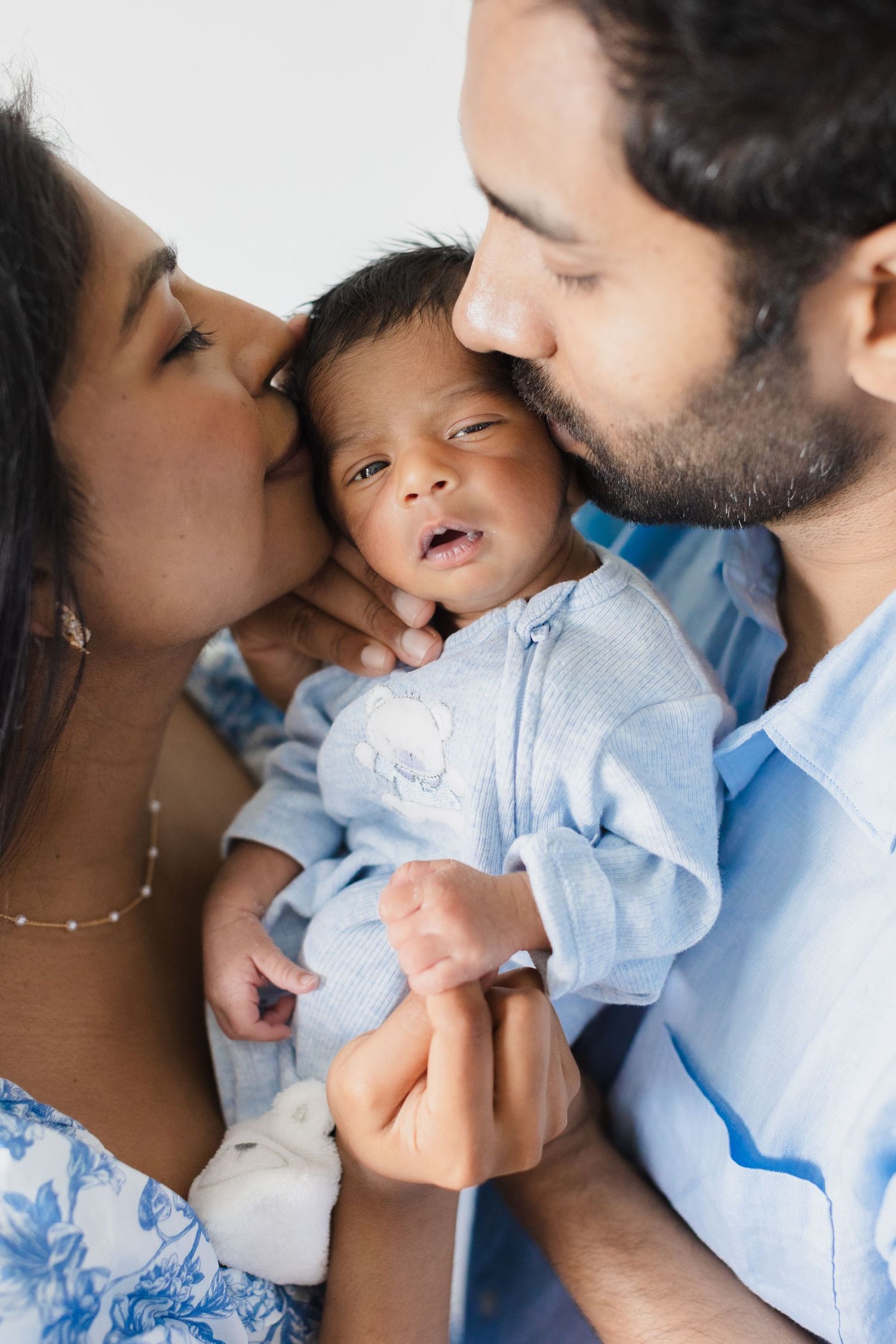 Mother and father kissing their newborn baby dressed in blue outfit.