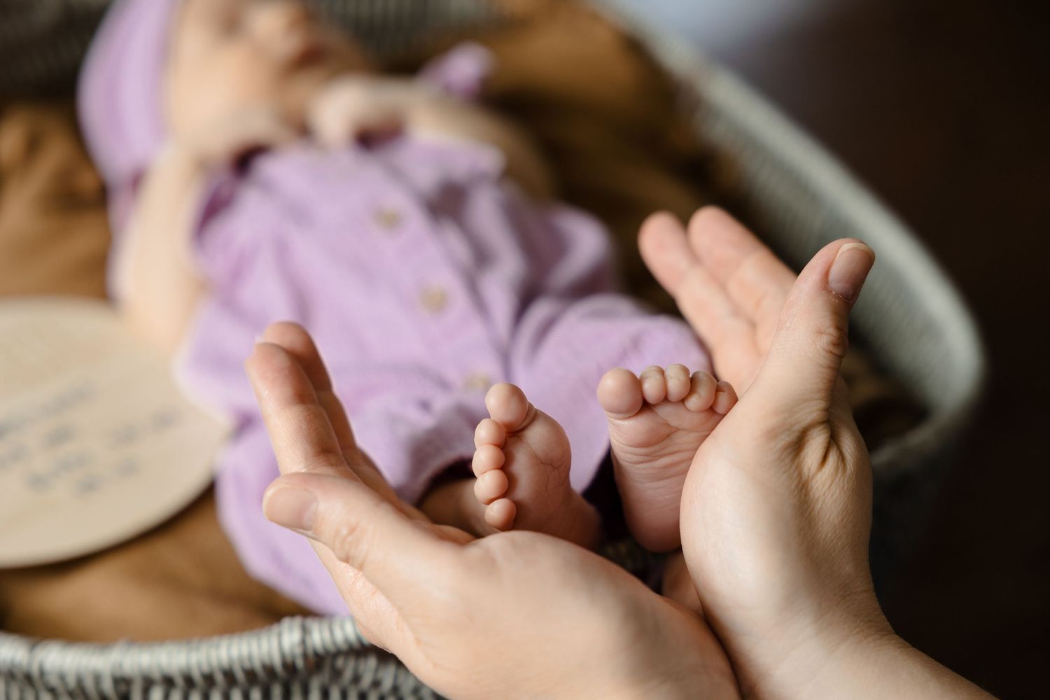 Adult hands gently hold tiny newborn baby feet dressed in purple outfit in wicker basket.