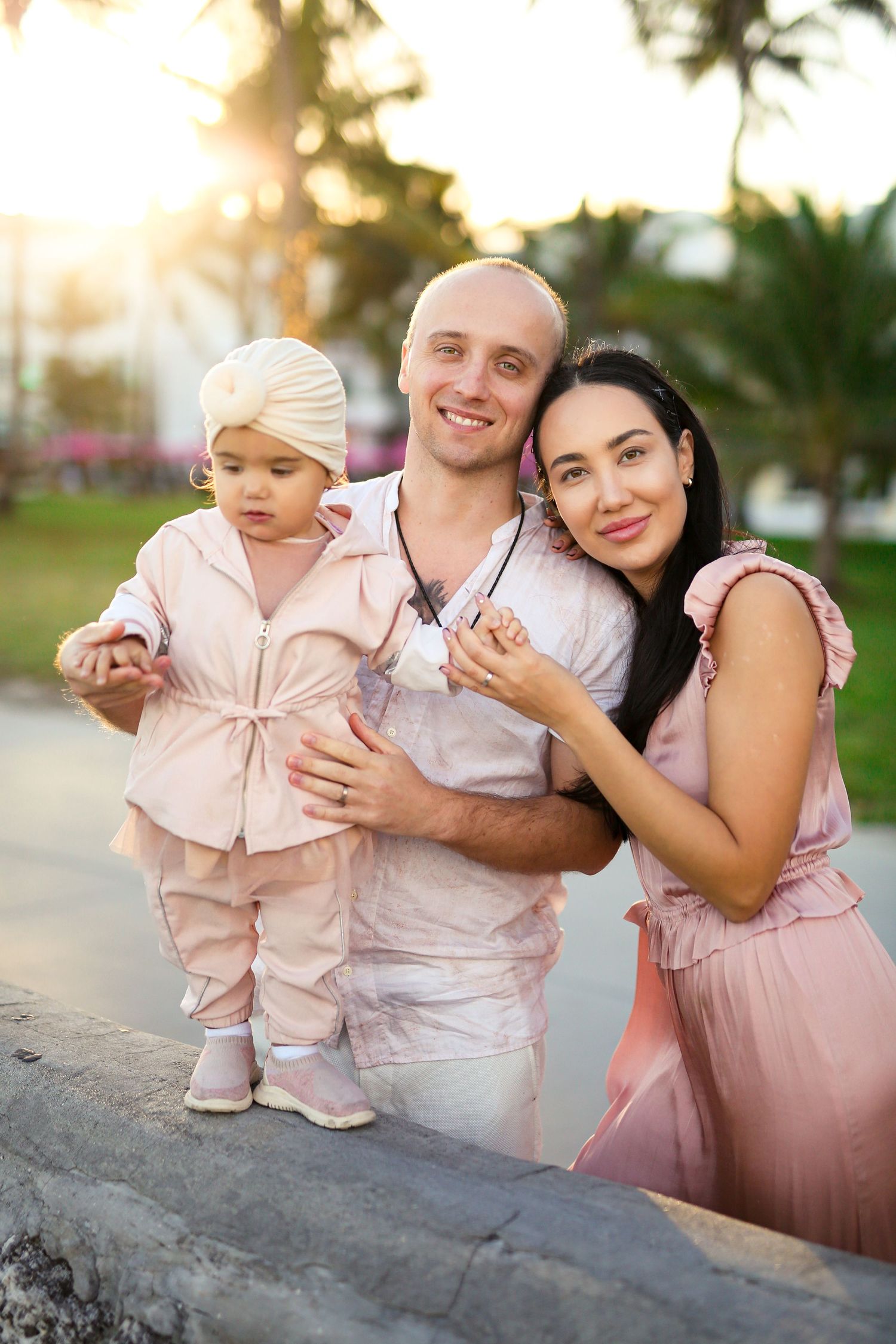 Smiling family of three in matching pink outfits posing at sunset near tropical palm trees.