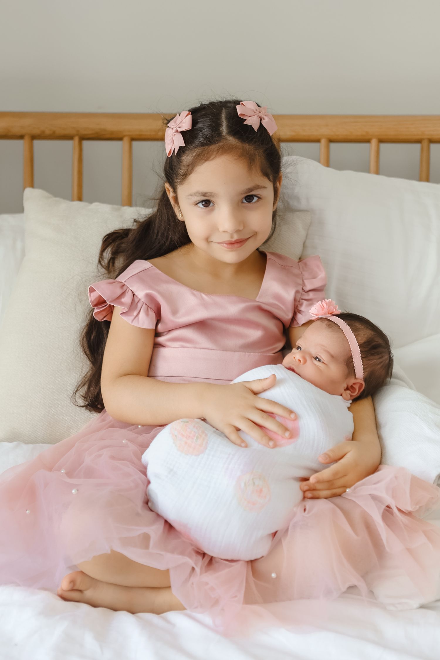 Young girl in pink dress holding swaddled newborn sibling on white bed.