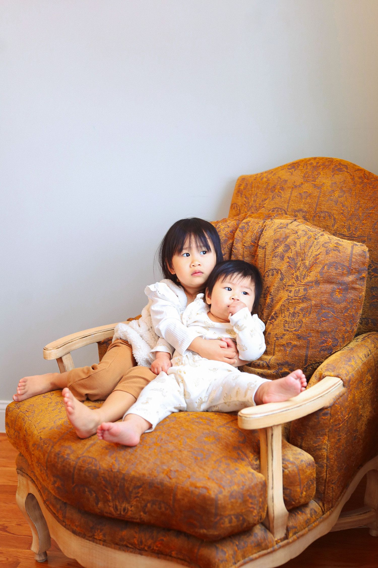 Two young Asian children sitting together in an ornate vintage armchair, looking at the camera.