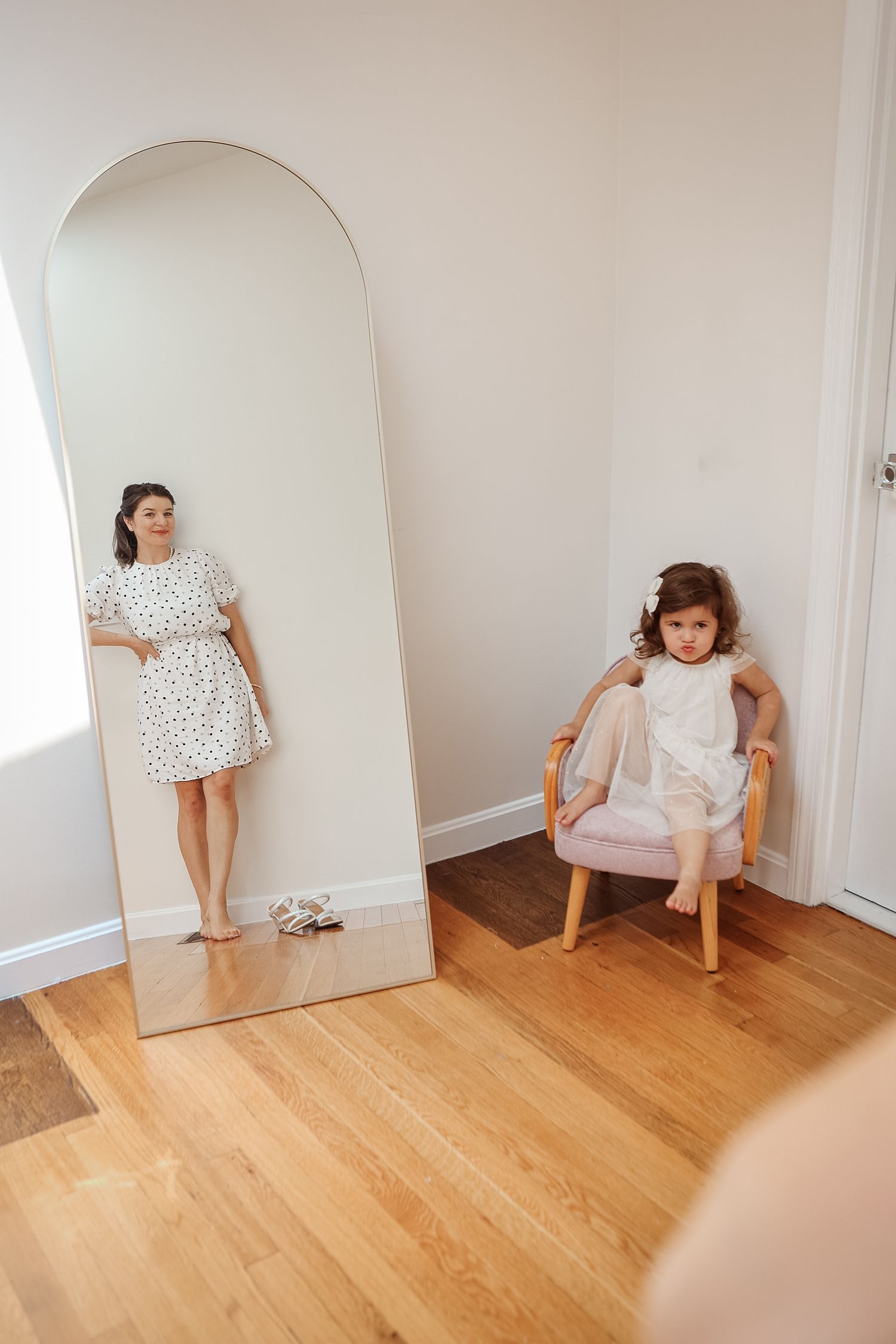 Young woman in white polka dot dress poses before tall arch mirror while little girl sits in pink chair nearby.