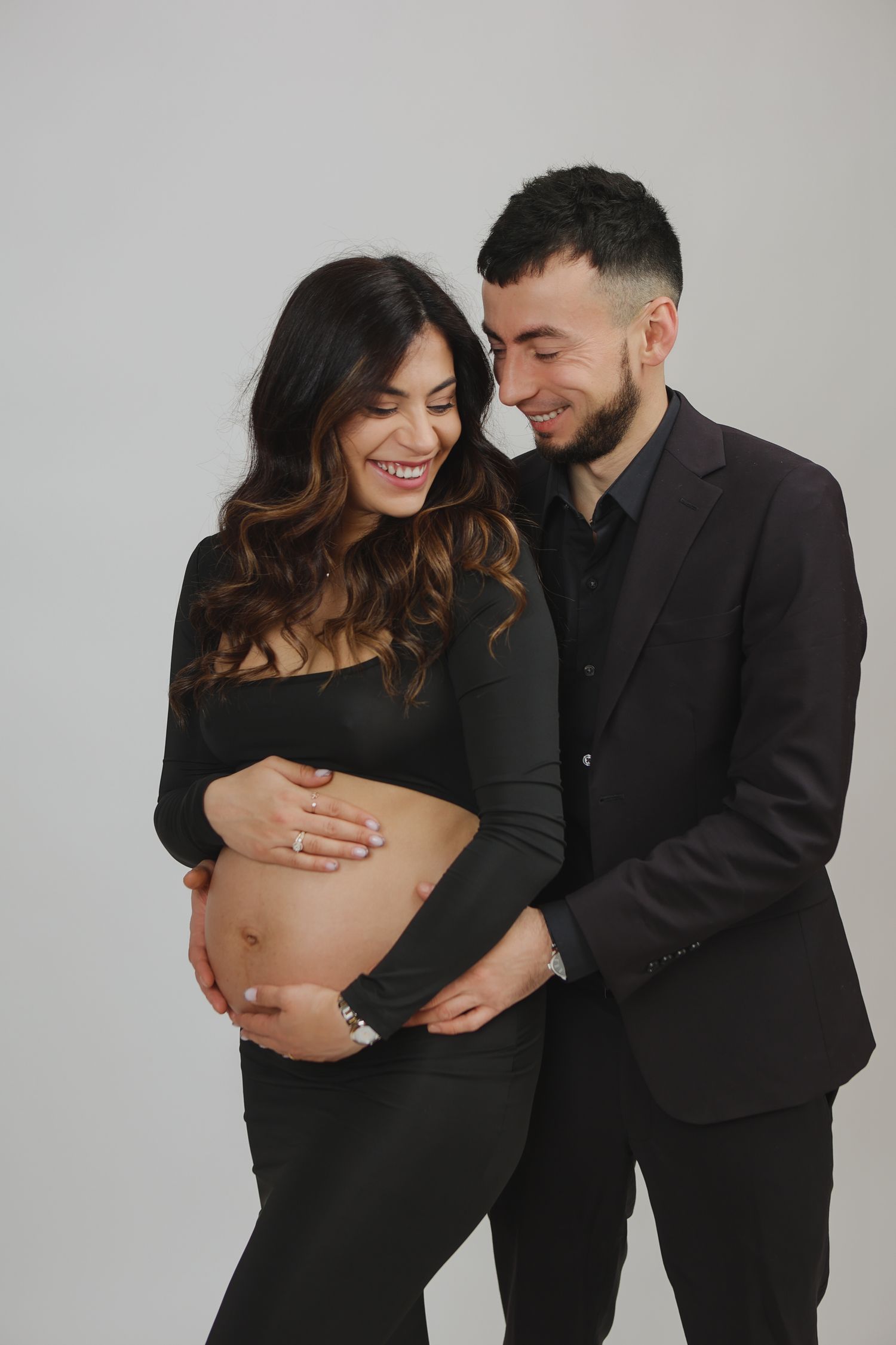 Happy expecting couple in matching black outfits smiling while cradling pregnant belly together.