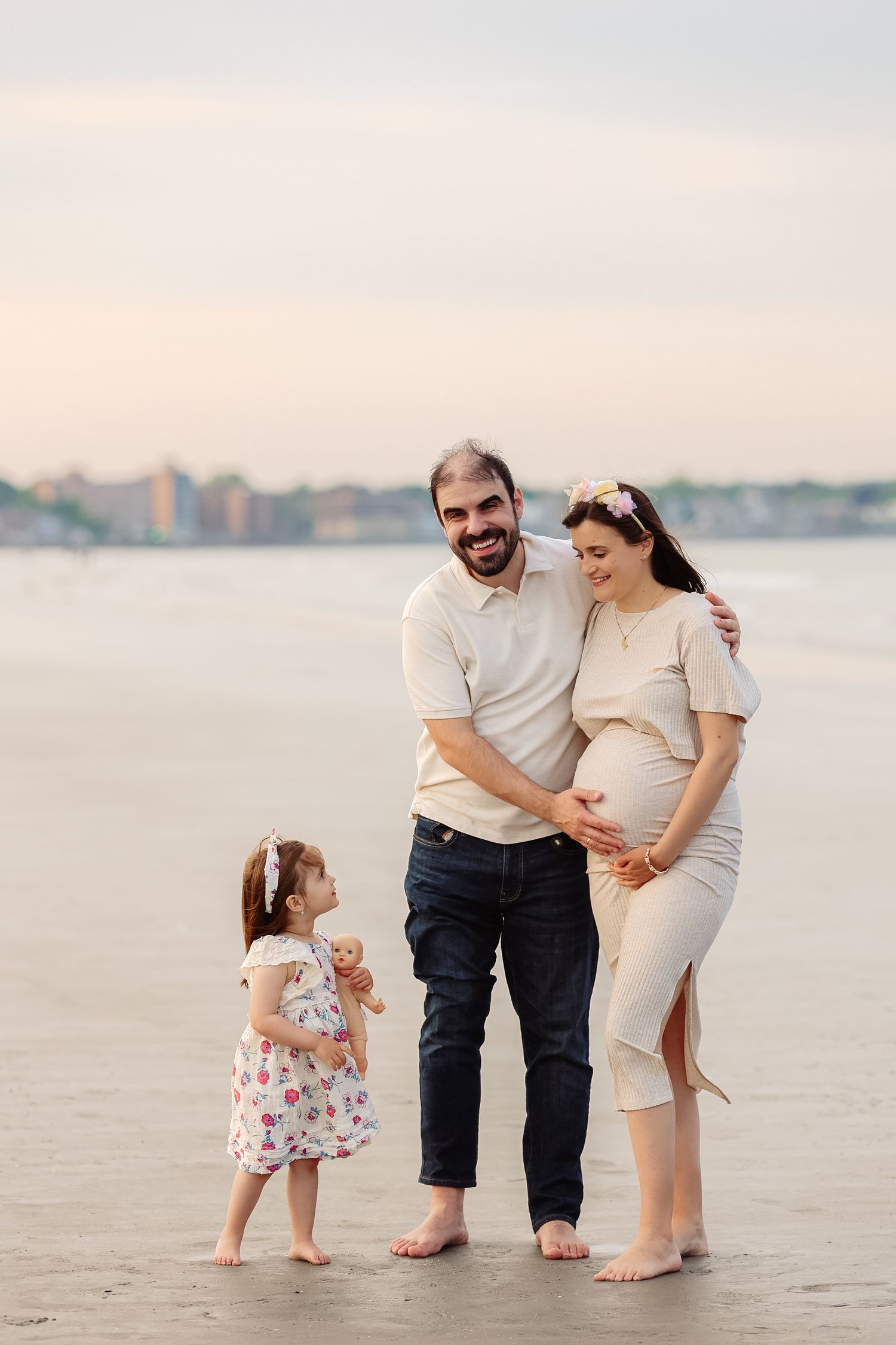 Happy family with pregnant mother, smiling father, and toddler daughter pose together on a wet beach at sunset.