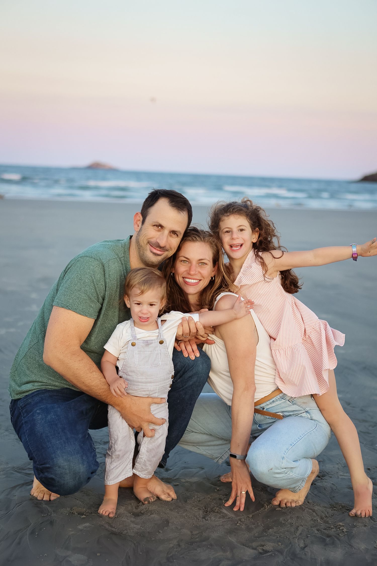 Smiling family of four crouching together on beach at sunset, daughter spreading arms wide.