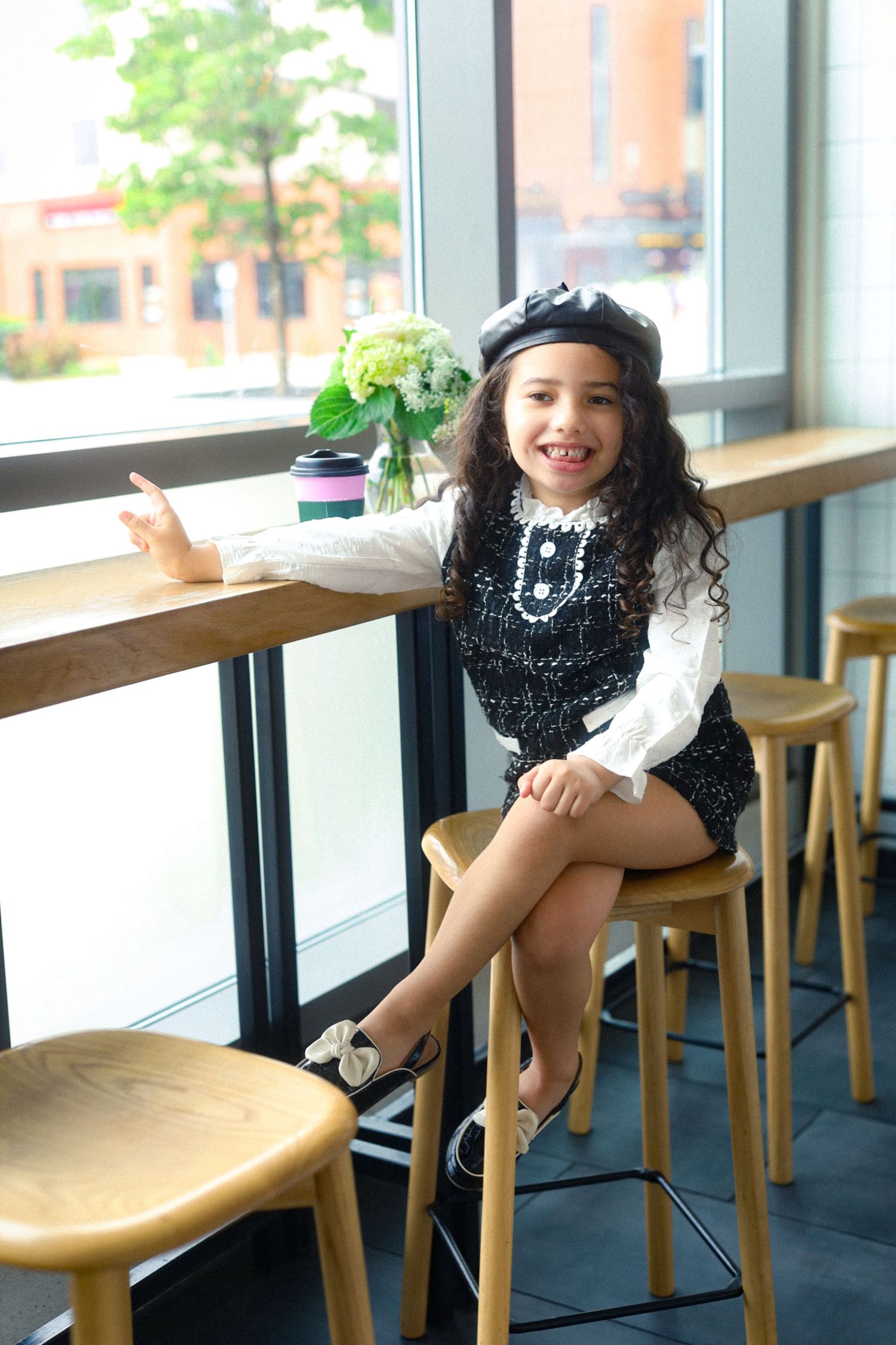 Stylish young girl in black beret and sequin outfit sitting on wooden stool by cafe window with flowers.