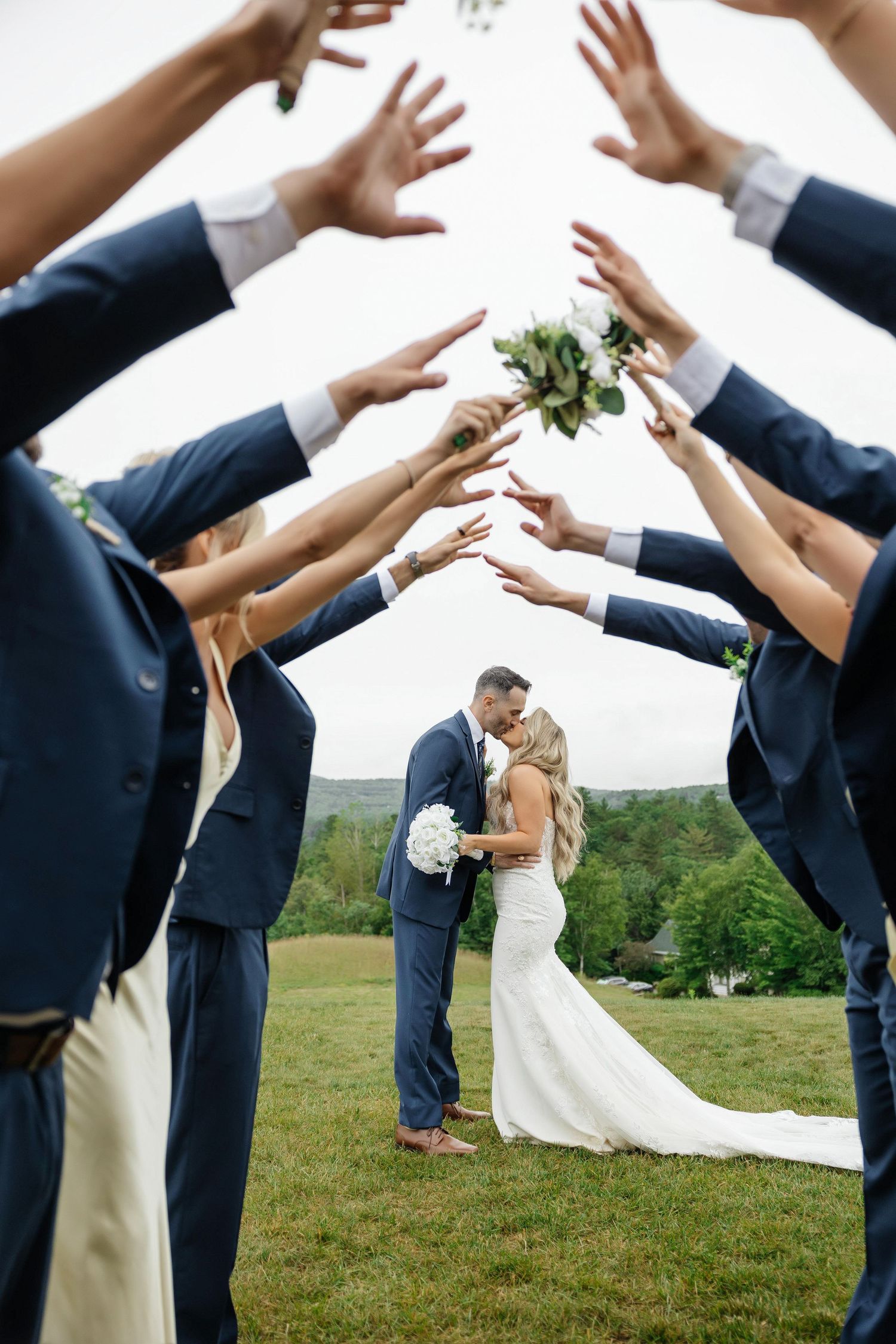 Wedding party creates arch with raised arms as bride and groom kiss at Owls Nest Resort in Thornton, New Hampshire, captured by Grand Photo