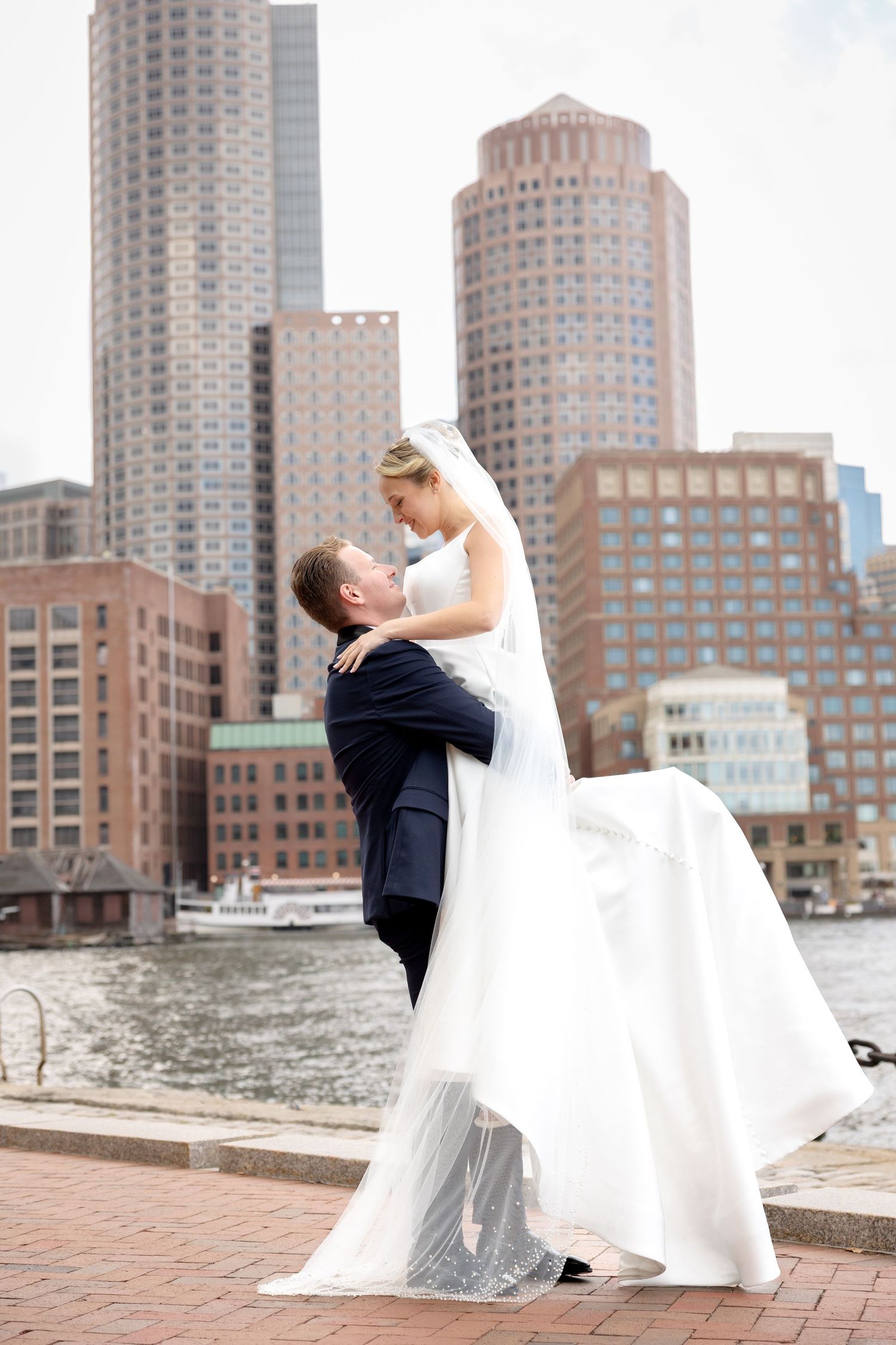 Groom lifts bride with Seaport Boston skyline background at Boston Harbor Hotel in Boston, Massachusetts, captured by Grand Photo