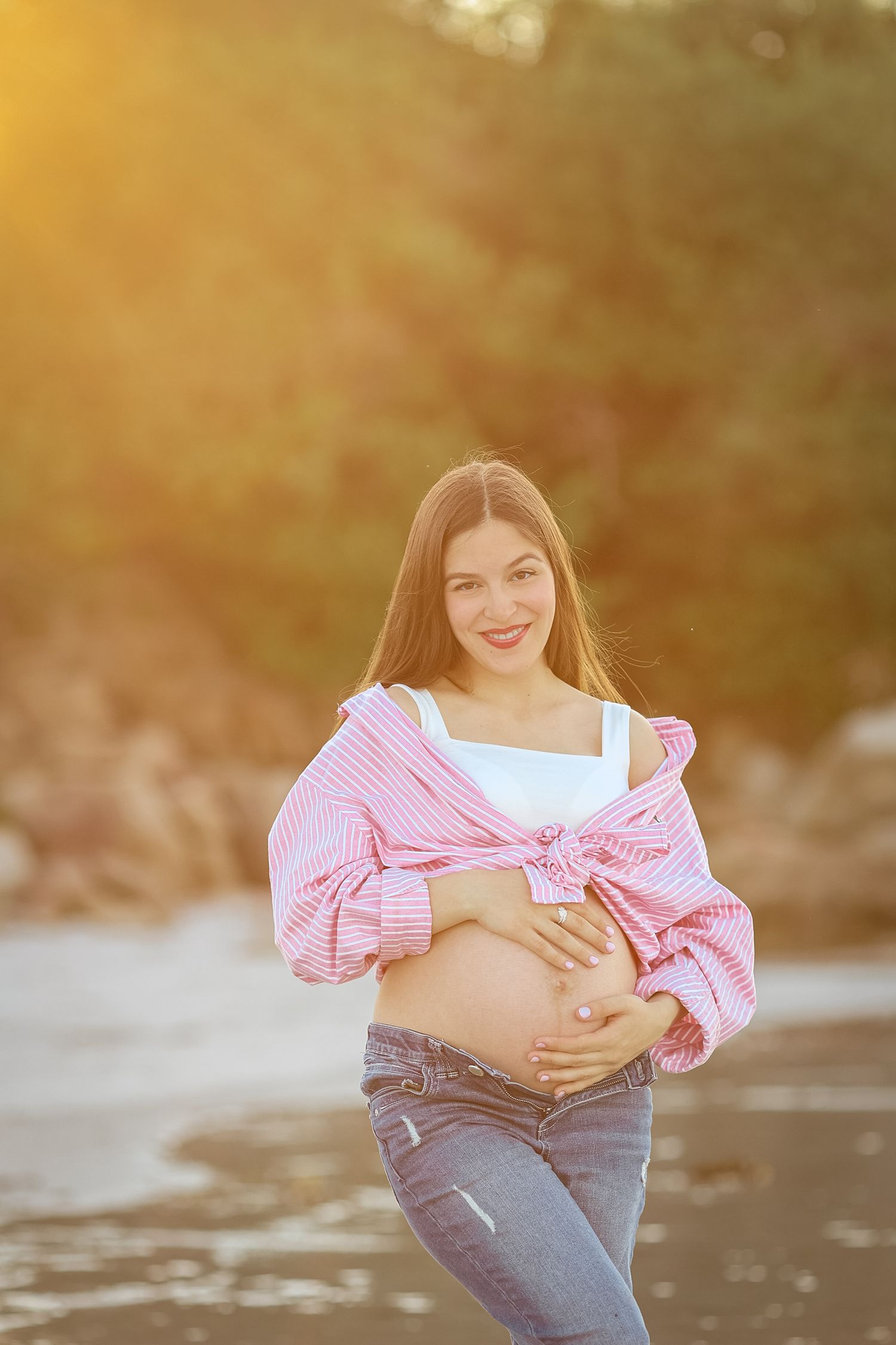 Smiling pregnant woman in pink striped shirt and jeans cradles her baby bump outdoors in golden sunset light.