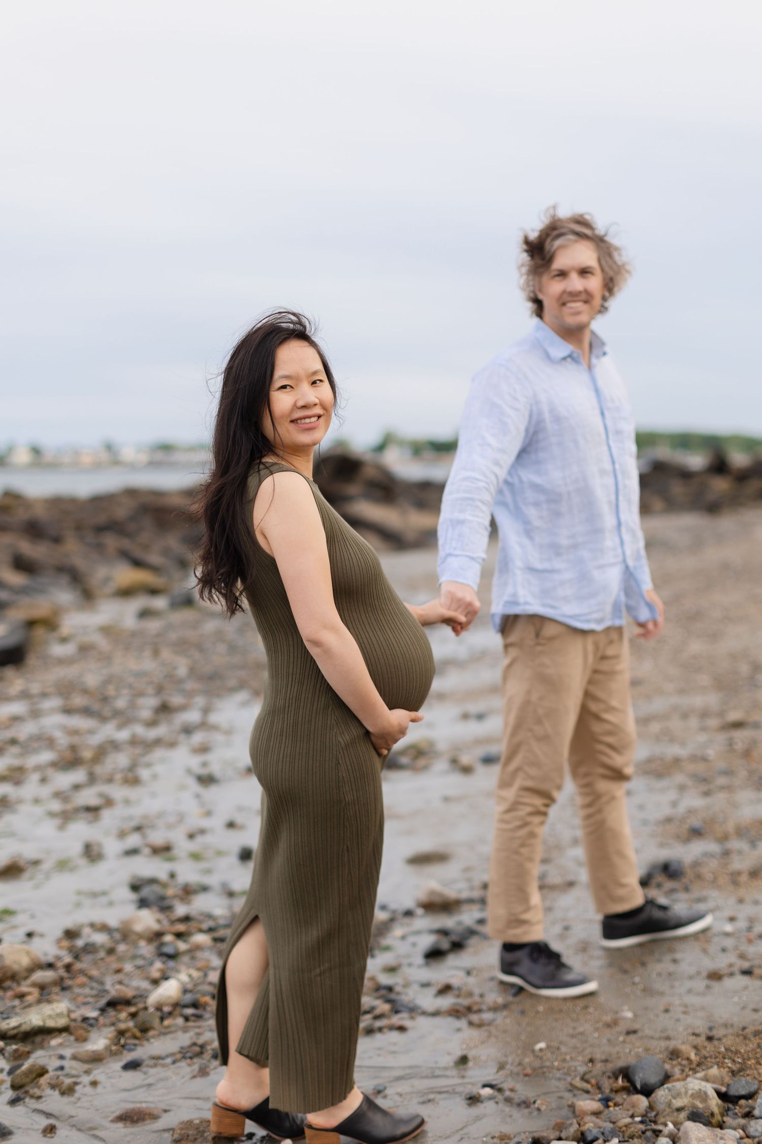 Pregnant woman in olive dress holds partners hand while smiling on rocky beach during maternity photoshoot.