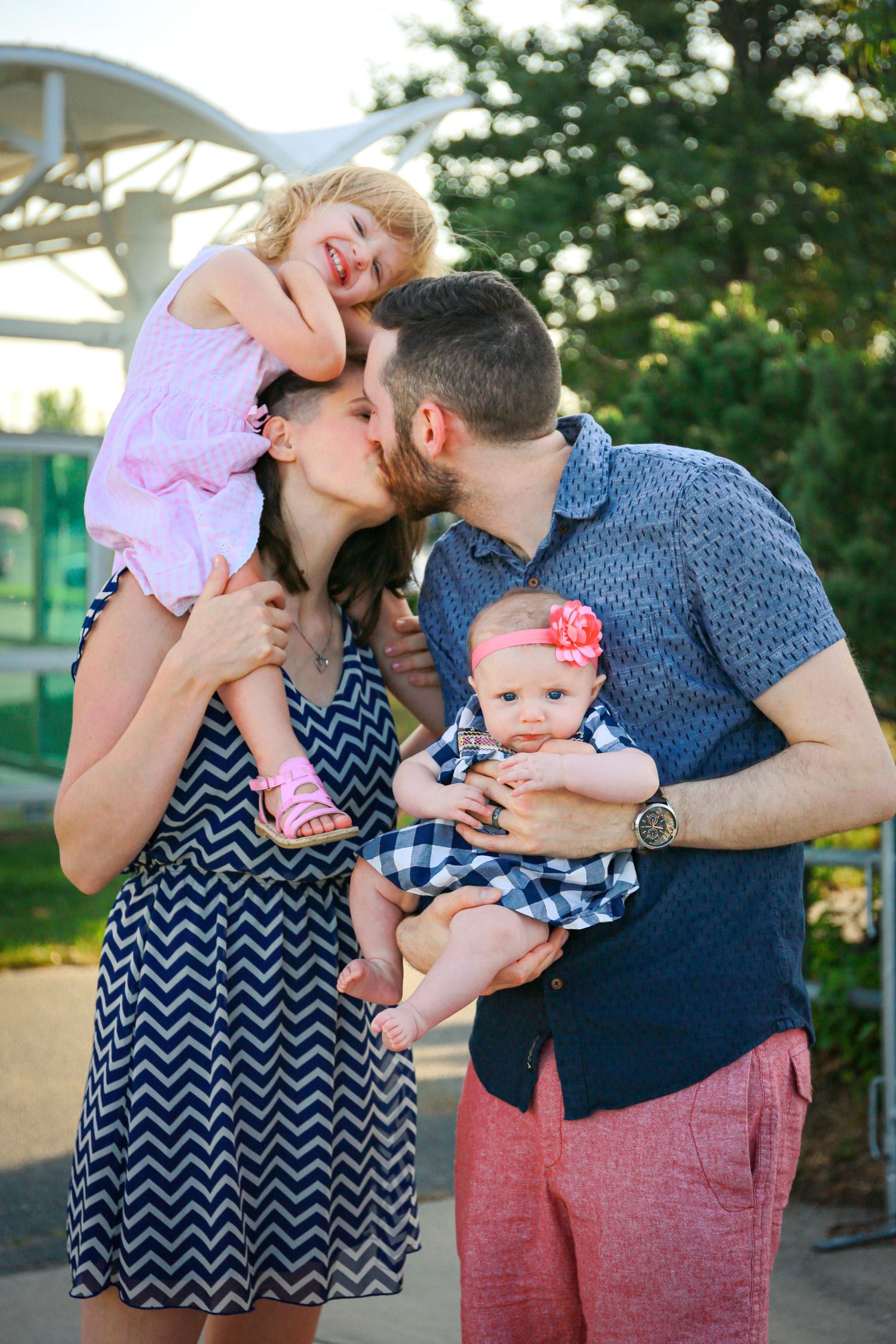Couple kissing outdoors while holding two young daughters, one on shoulders and one in arms.