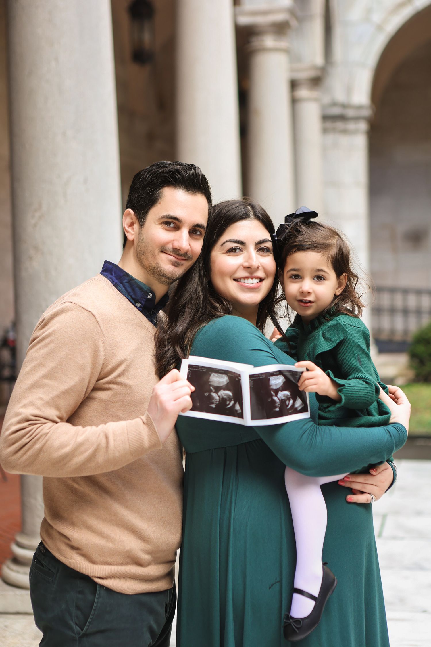 Happy family holding ultrasound photos during maternity announcement session at historic building.