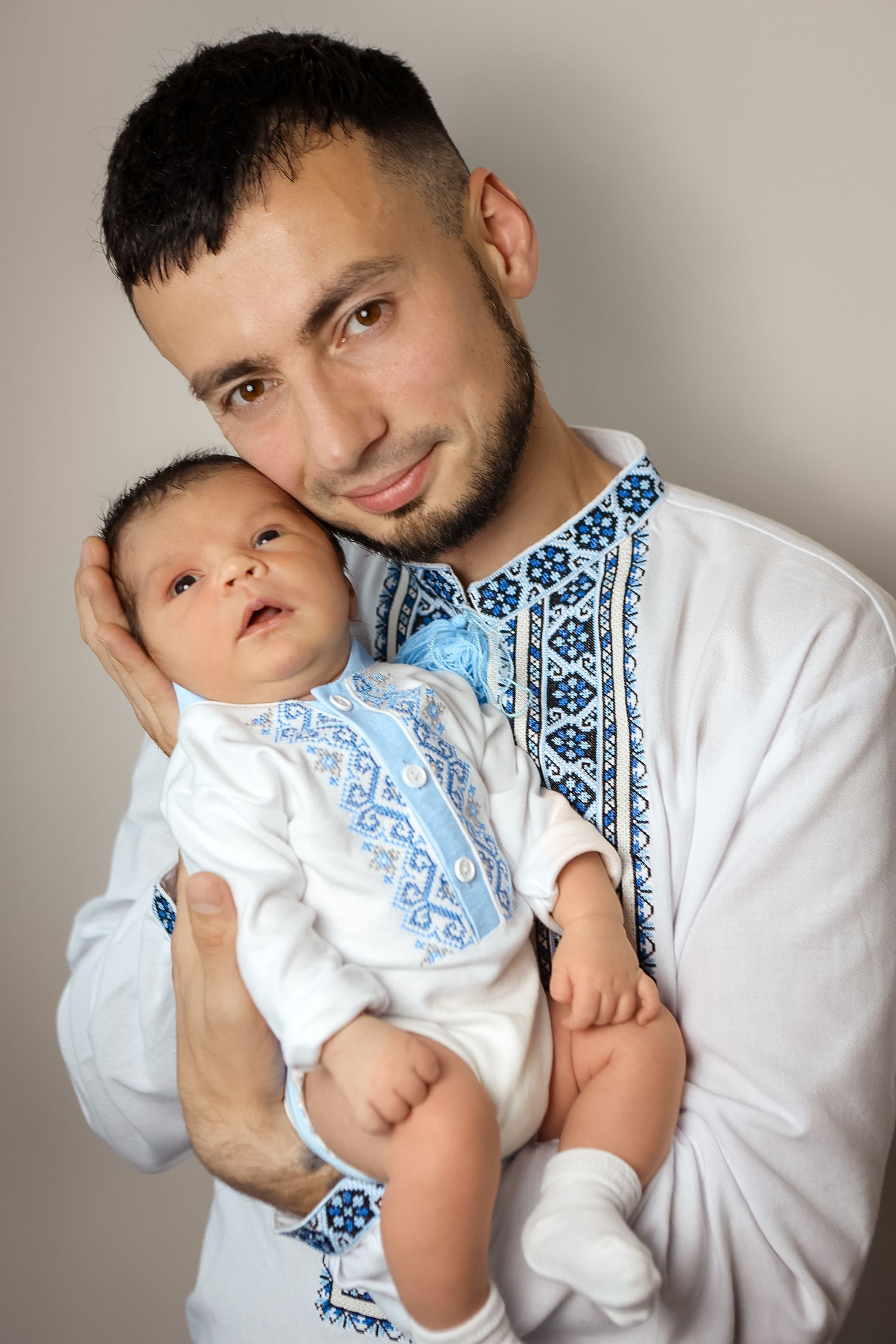 Young father in traditional Ukrainian embroidered vyshyvanka shirt holds newborn baby against neutral background.