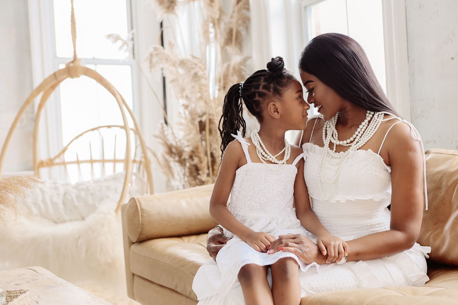 Mother and daughter in matching white dresses with pearl necklaces sitting together on beige sofa.