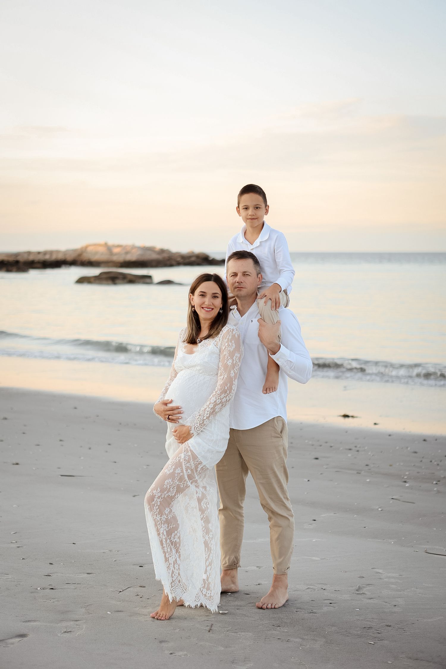 Family maternity portrait on beach at sunset with pregnant mother in white lace dress.