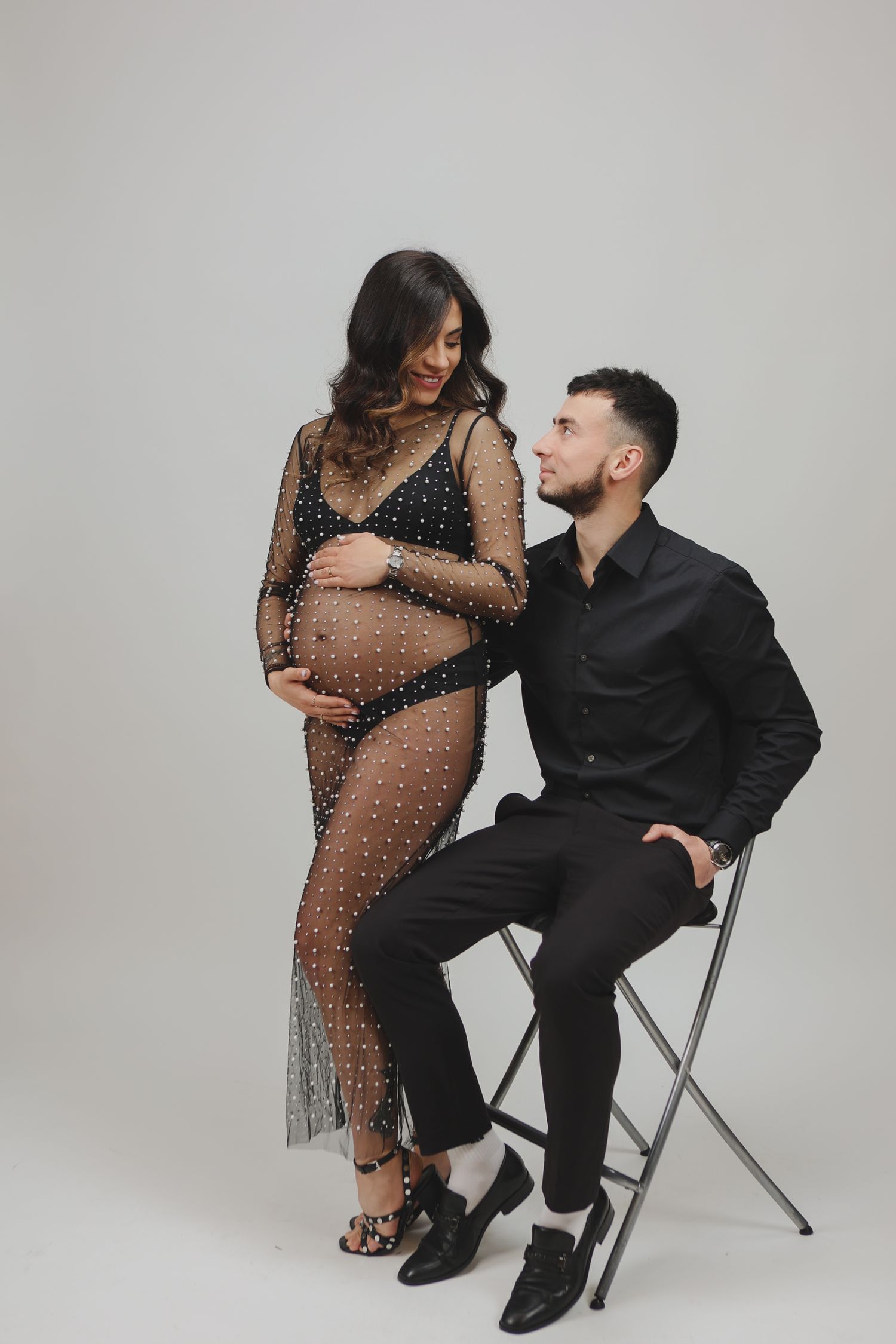 Pregnant woman in sheer black beaded dress standing beside seated partner in black shirt.