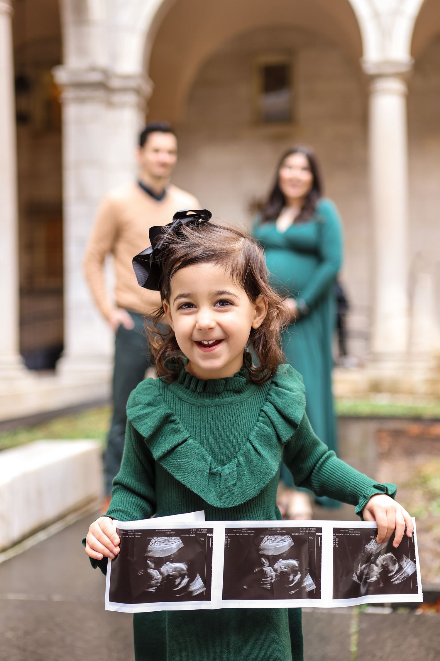 Excited little girl in green dress holding ultrasound photos with pregnant parents blurred in background.