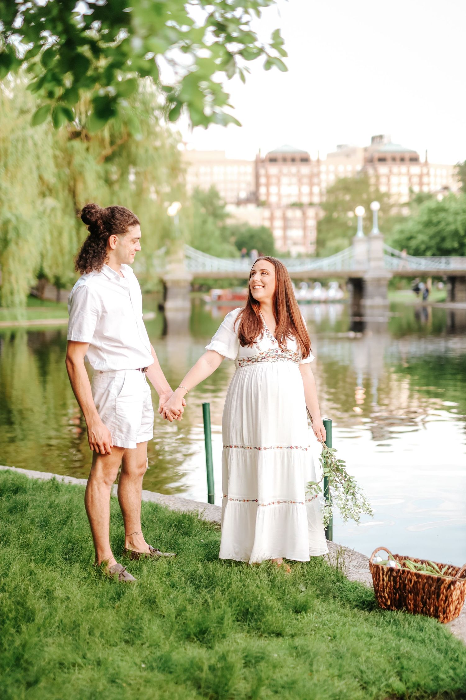 Pregnant woman in white dress holds wildflowers while couple walks hand in hand by park pond.