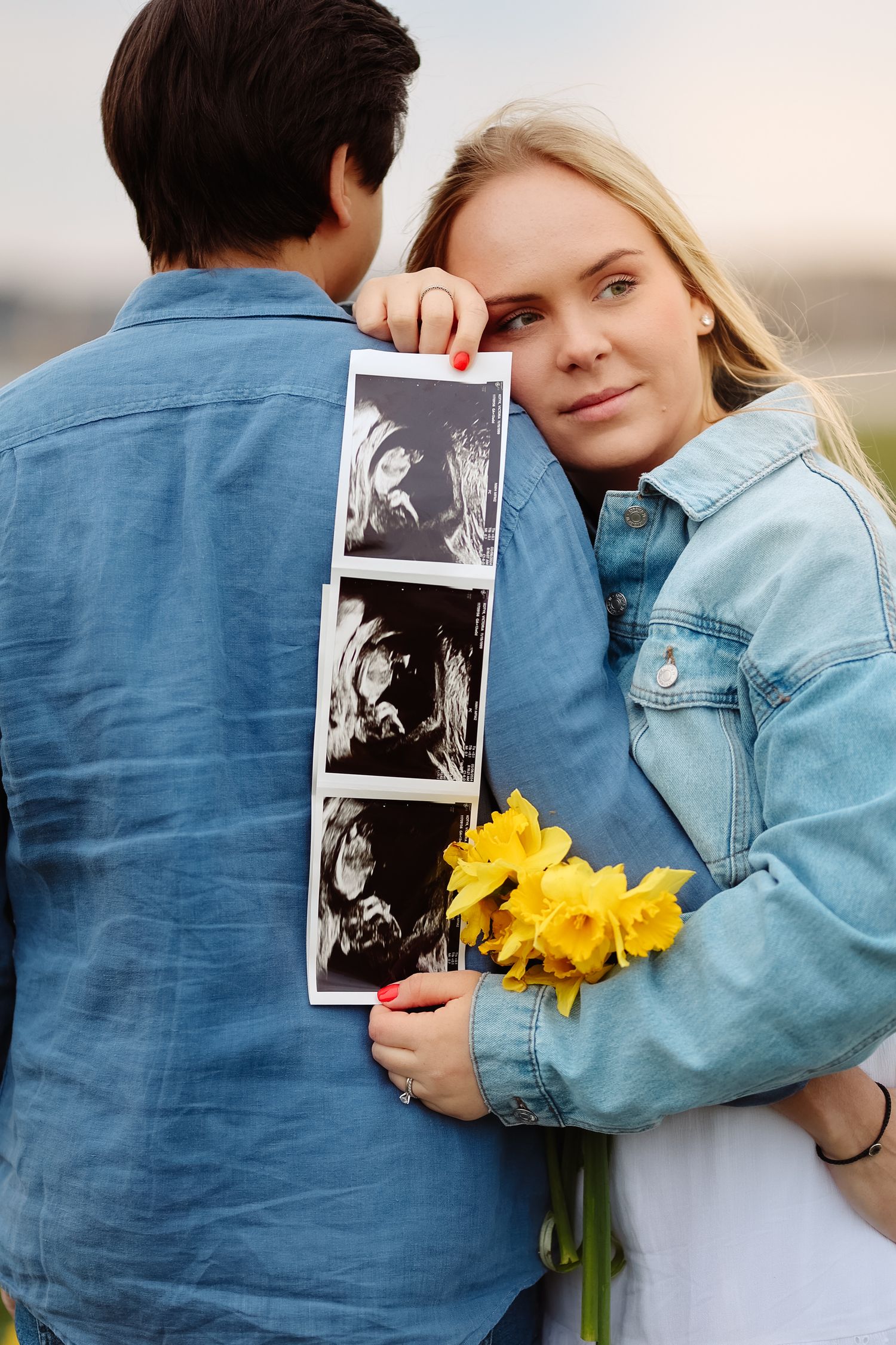 Pregnant blonde woman leaning on partner showing ultrasound images while holding yellow daffodils outdoors.